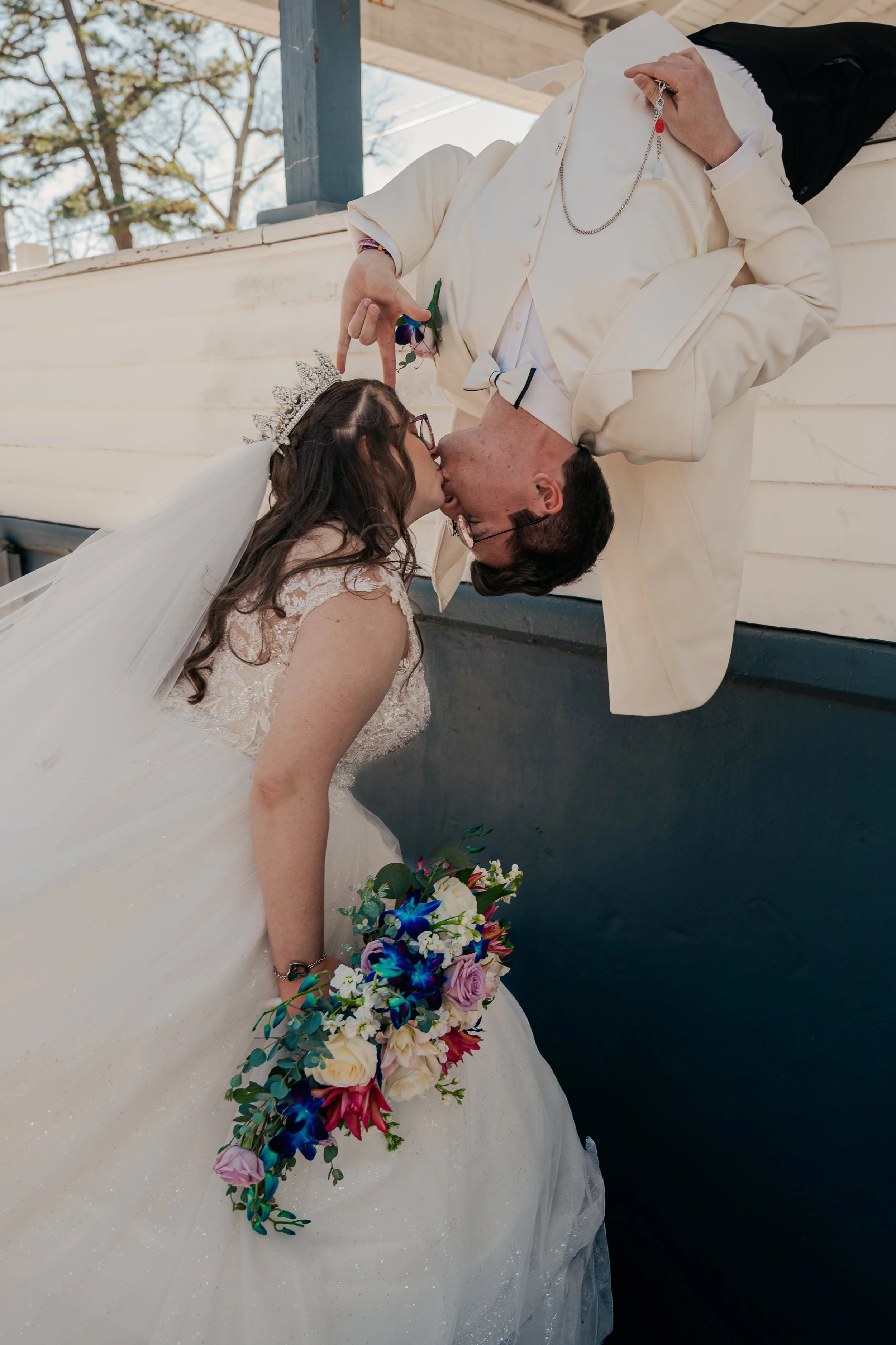 A newlywed couple sharing a kiss by a body of water, with the bride holding a colorful bouquet and wearing a wedding dress and tiara, and the groom in a cream tuxedo. Johnstown Pennsylvania, Pittsburgh, New Jersey, elopement Photographer, elope