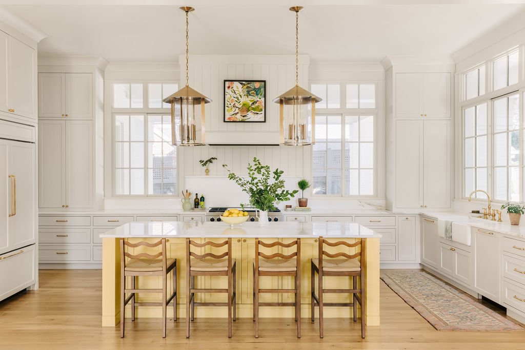 Modern kitchen with white cabinetry, large windows, and a central island with wooden chairs. Features brass pendant lights, a small plant, and a bowl of lemons on the counter.
