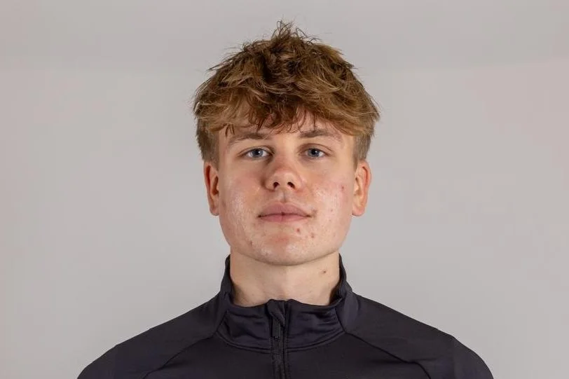 A young man with blonde, slightly tousled hair wearing a black zip-up jacket, neutral expression, against a plain grey background.