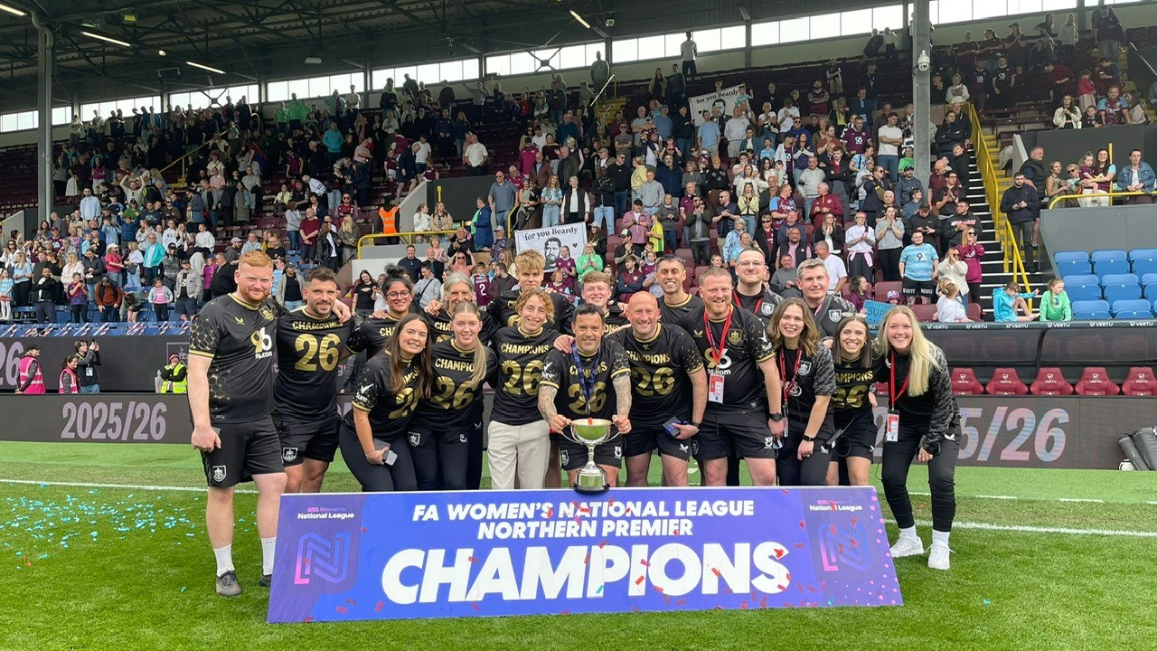 A group of women and men soccer coaches and staff celebrating with a trophy and a large banner that reads "CHAMPIONS" after winning a match, with a crowd of spectators in the background inside a stadium.