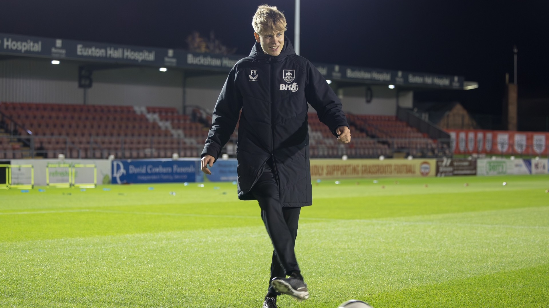 Young male soccer coach or trainer with blond hair wearing a black jacket, black pants, and black shoes, smiling while walking on a soccer field at night. The stadium seats are visible in the background, along with some training equipment on the field.