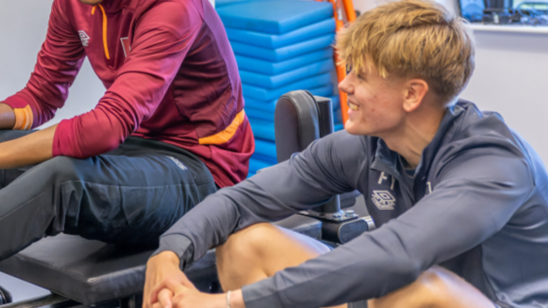 Two young men sitting on exercise benches in a fitness centre, smiling and having a conversation.