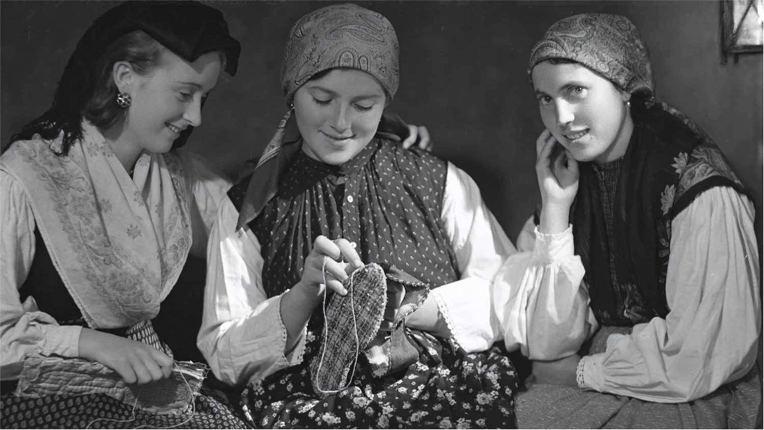 Three Friulian women in traditional clothing hand-stitching velvet slippers, preserving artisanal skills passed down generations.