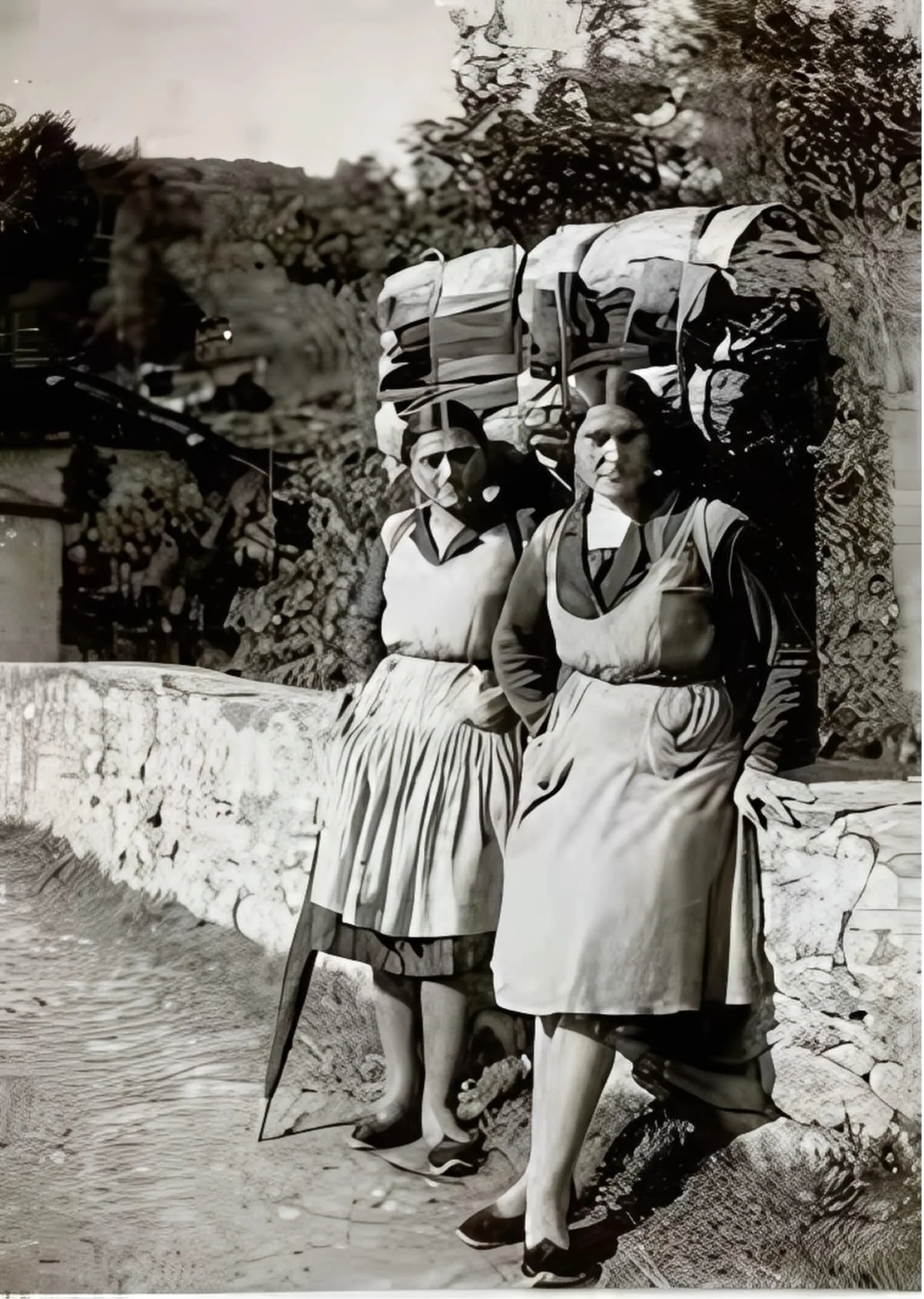 Historical image of two Friulian women carrying heavy loads and wearing friulana shoes, representing how this footwear was by peasants in their labor.