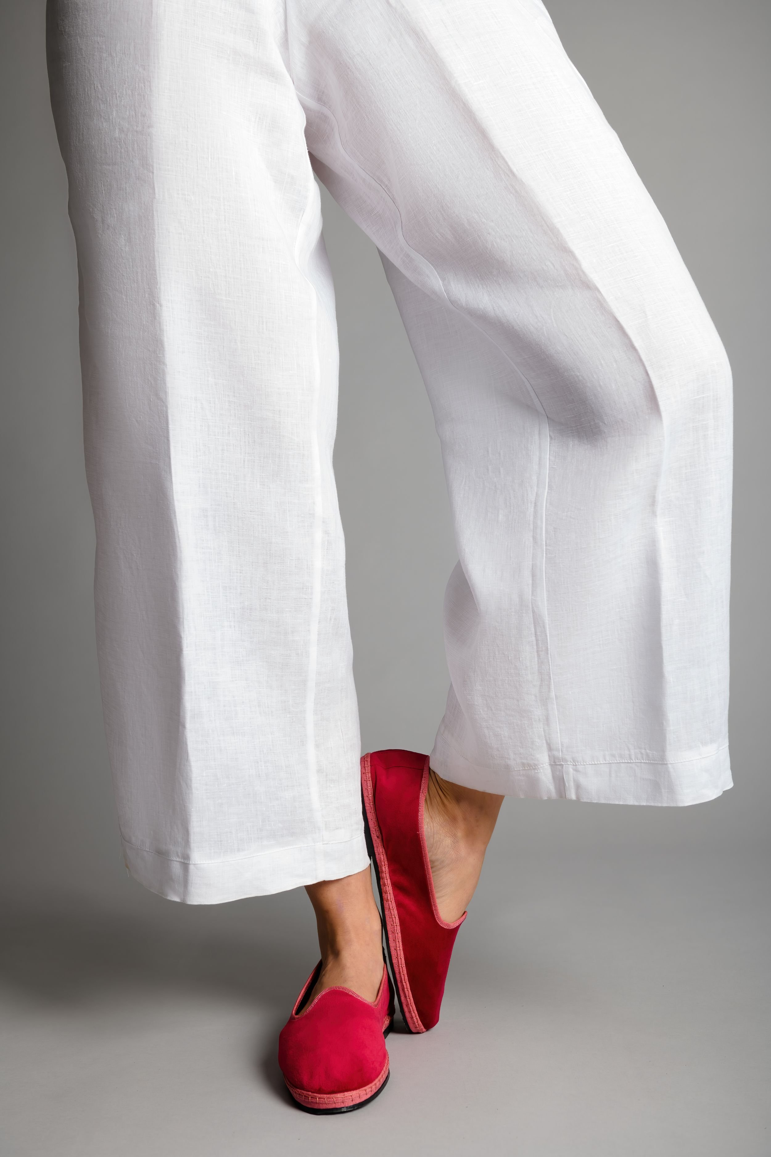 Model in white trousers with one foot lifted and crossed, wearing rose velvet Gioia slippers
