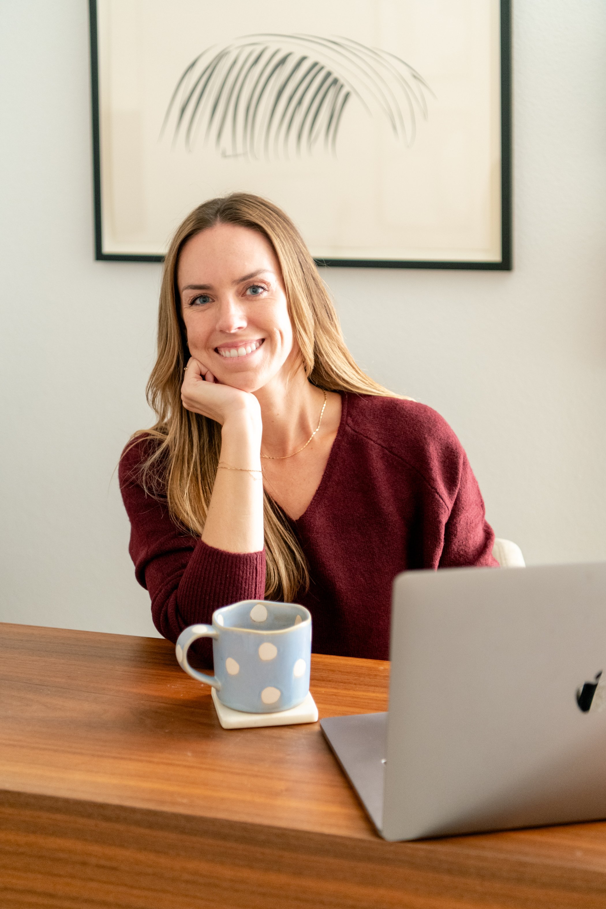 A woman with long brown hair, wearing a maroon sweater, sitting at a wooden table in front of a laptop and a polka-dotted mug, smiling at the camera against a background with a framed abstract line art on the wall.
