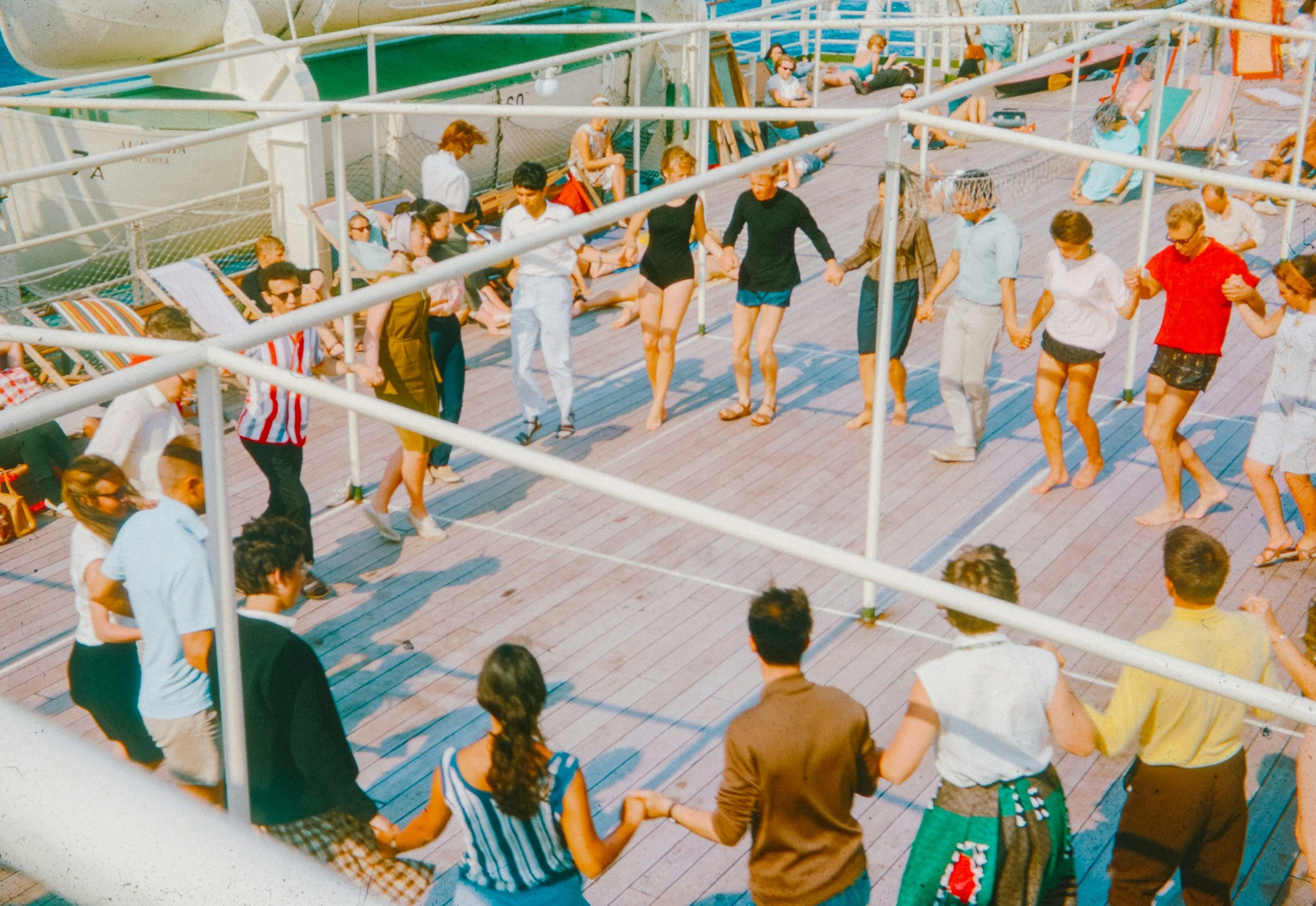 People holding hands and dancing in a circle on a ship's deck, with others relaxing on lounge chairs in the background.