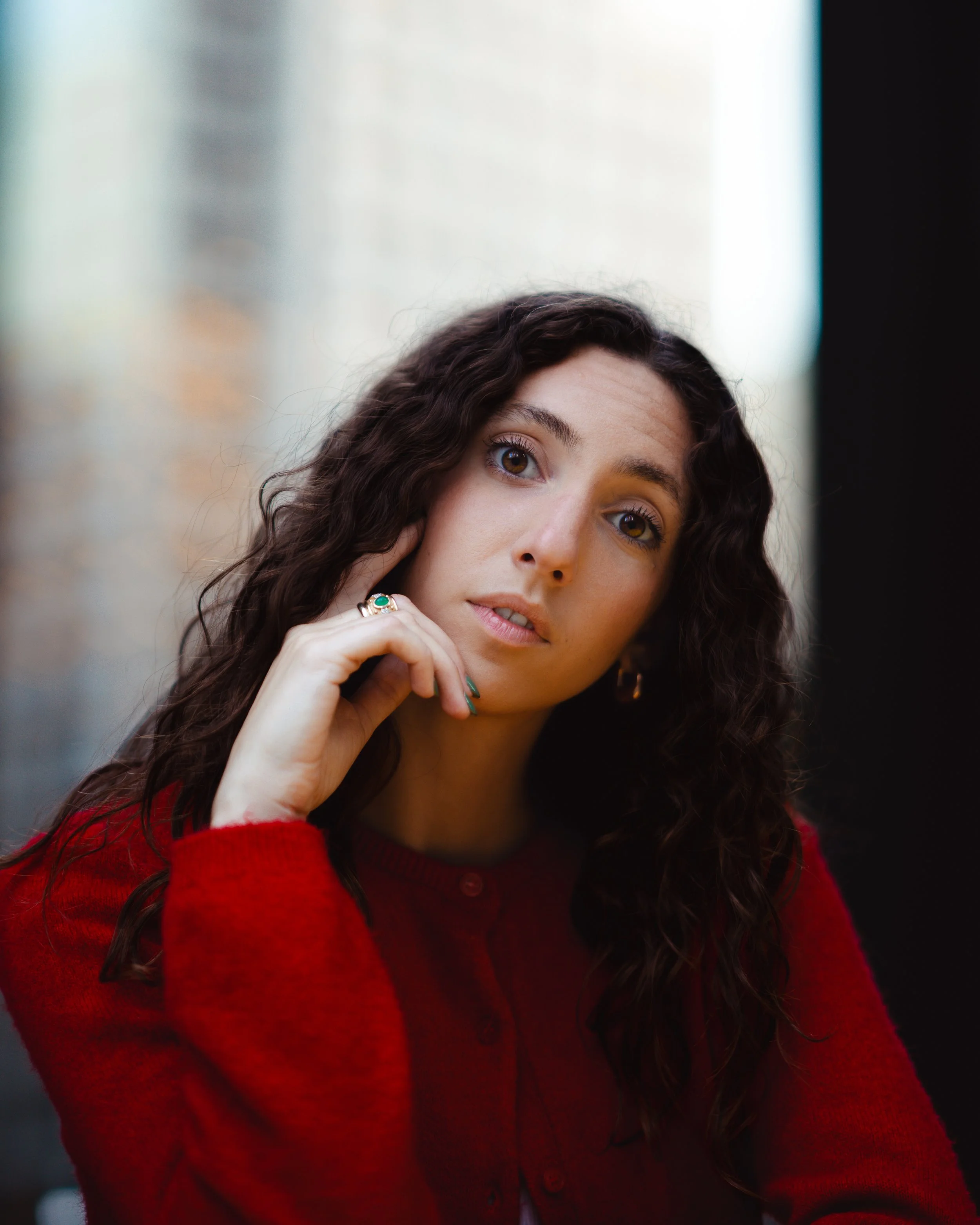 A woman with long, curly dark hair, wearing a red sweater, looking directly at the camera with her head slightly tilted to her right, resting her chin on her left hand, in an urban setting with blurred buildings in the background.
