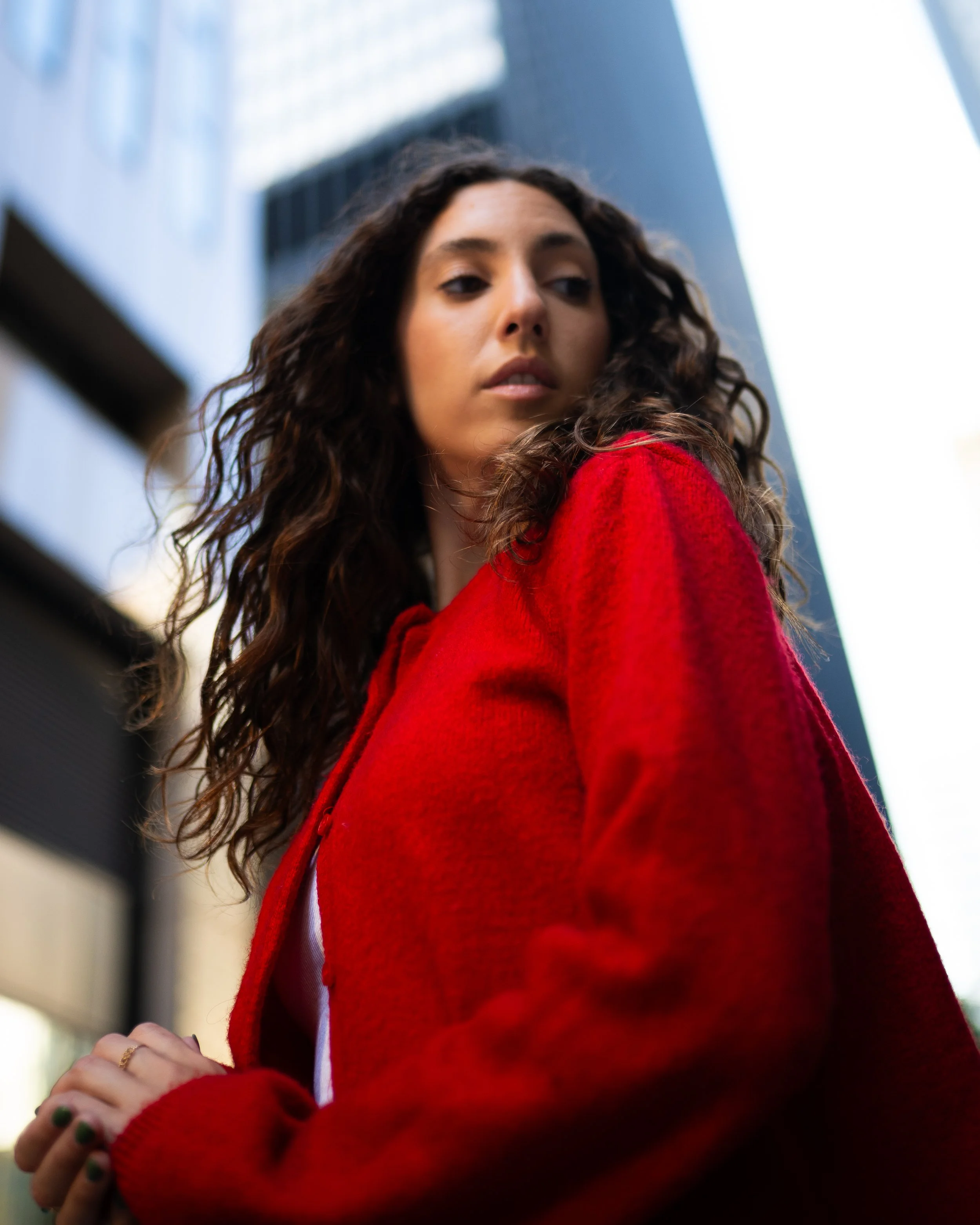 A woman with curly brown hair wearing a red coat, looking down over her shoulder with city buildings in the background.