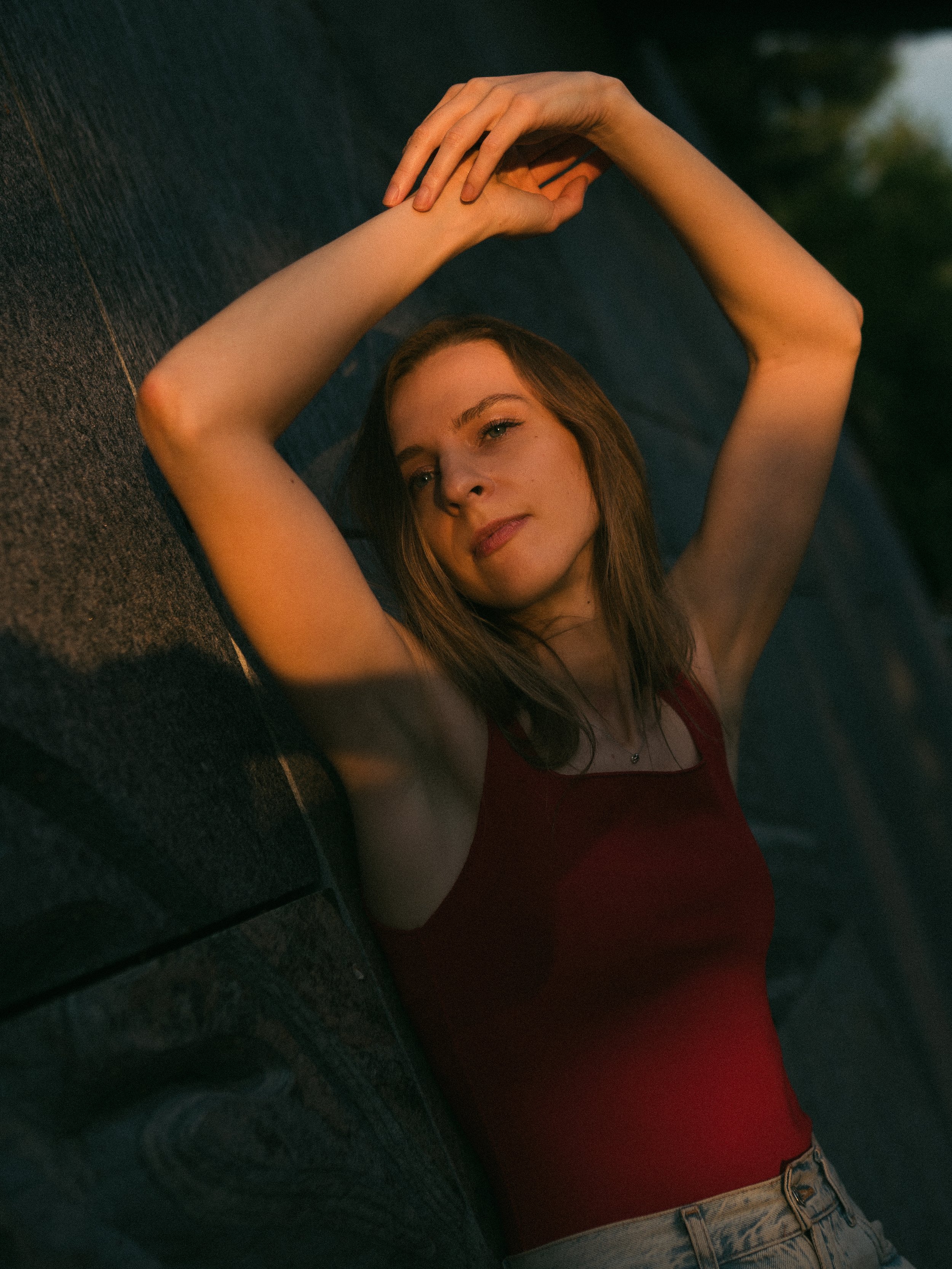 A young woman with red hair lying on the ground, wearing a red top and jeans, with arms raised above her head and looking thoughtfully into the distance during sunset.