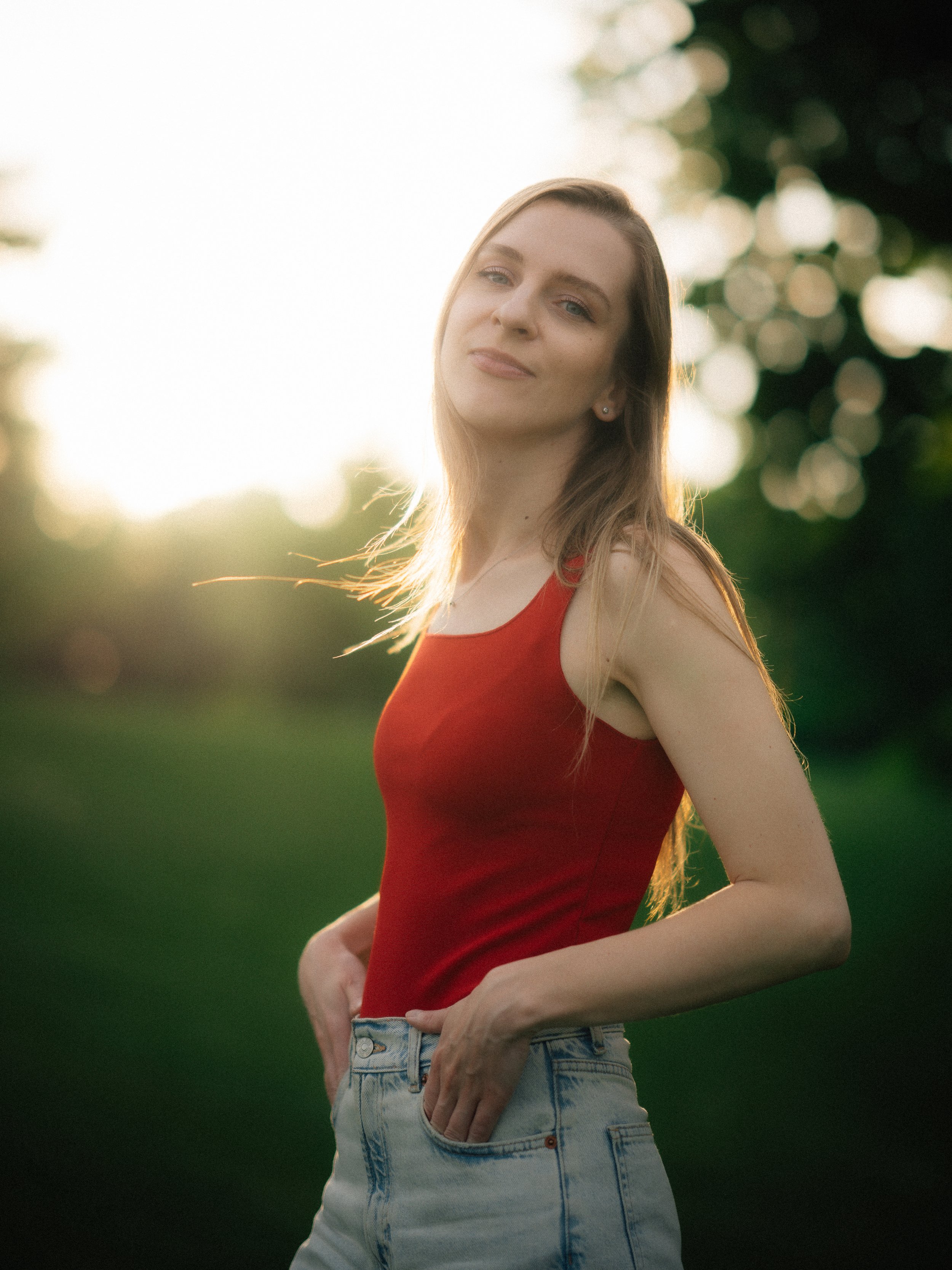 A woman with long, light brown hair wearing a red tank top and light blue jeans standing outdoors during sunset.