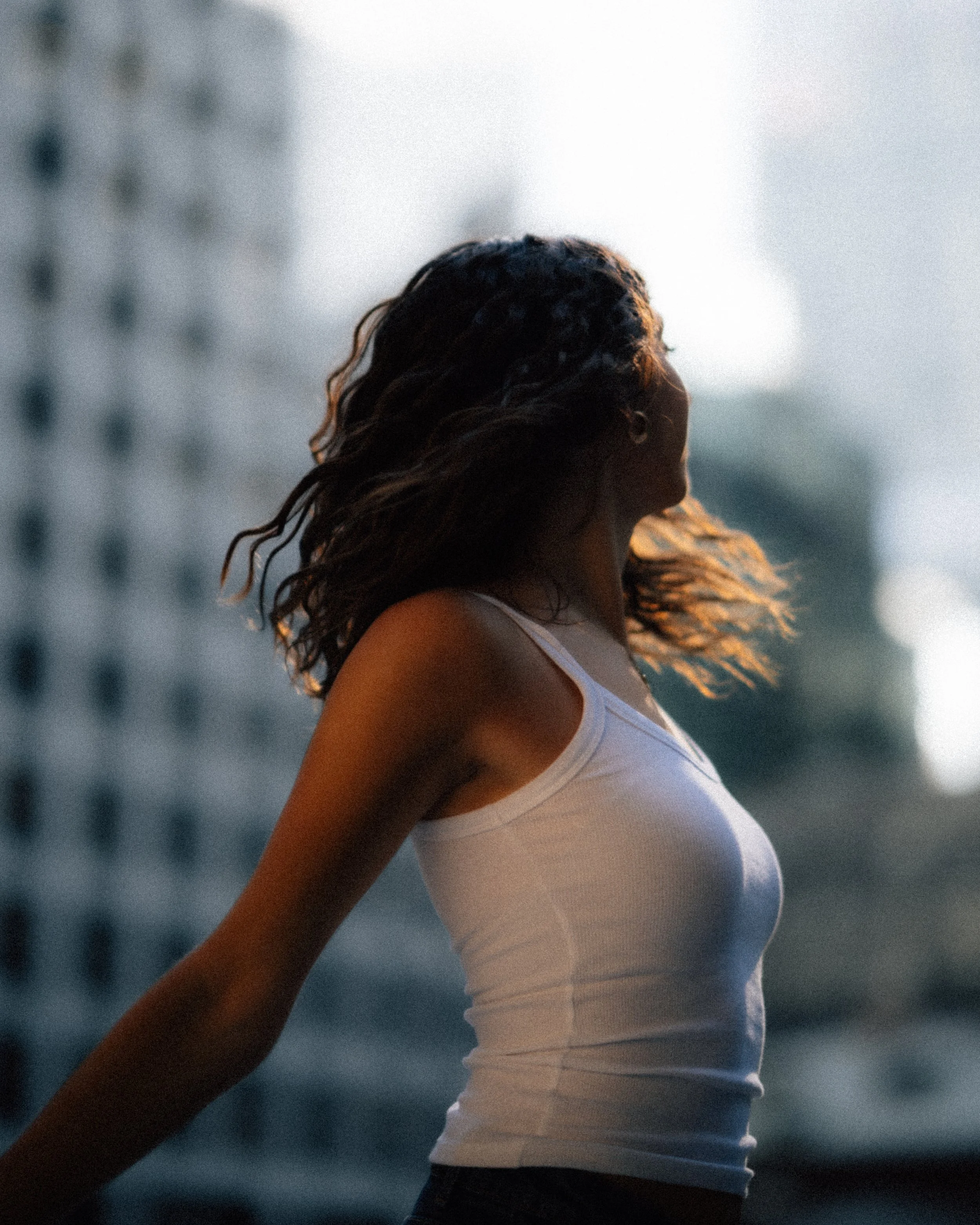 A woman with curly hair wearing a white tank top standing outdoors in front of a blurred cityscape background.