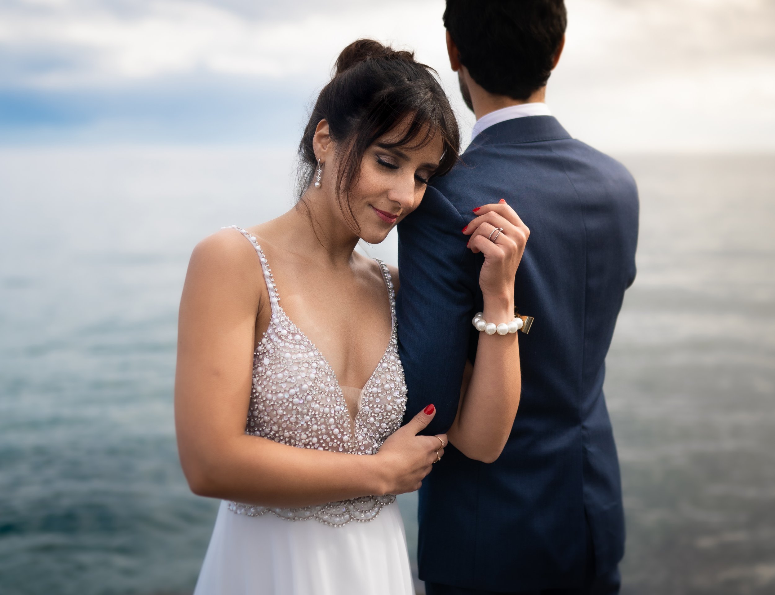 A woman in a wedding dress leans on a man in a suit near the water, looking content with closed eyes.
