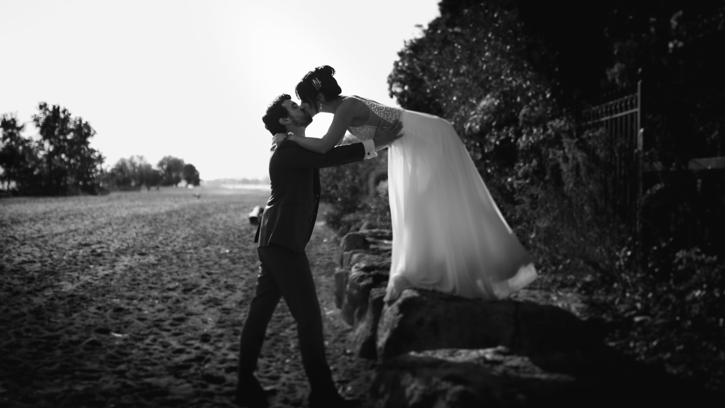 A black and white photo of a man lifting a woman in a wedding dress in a park, with trees and an open field in the background.