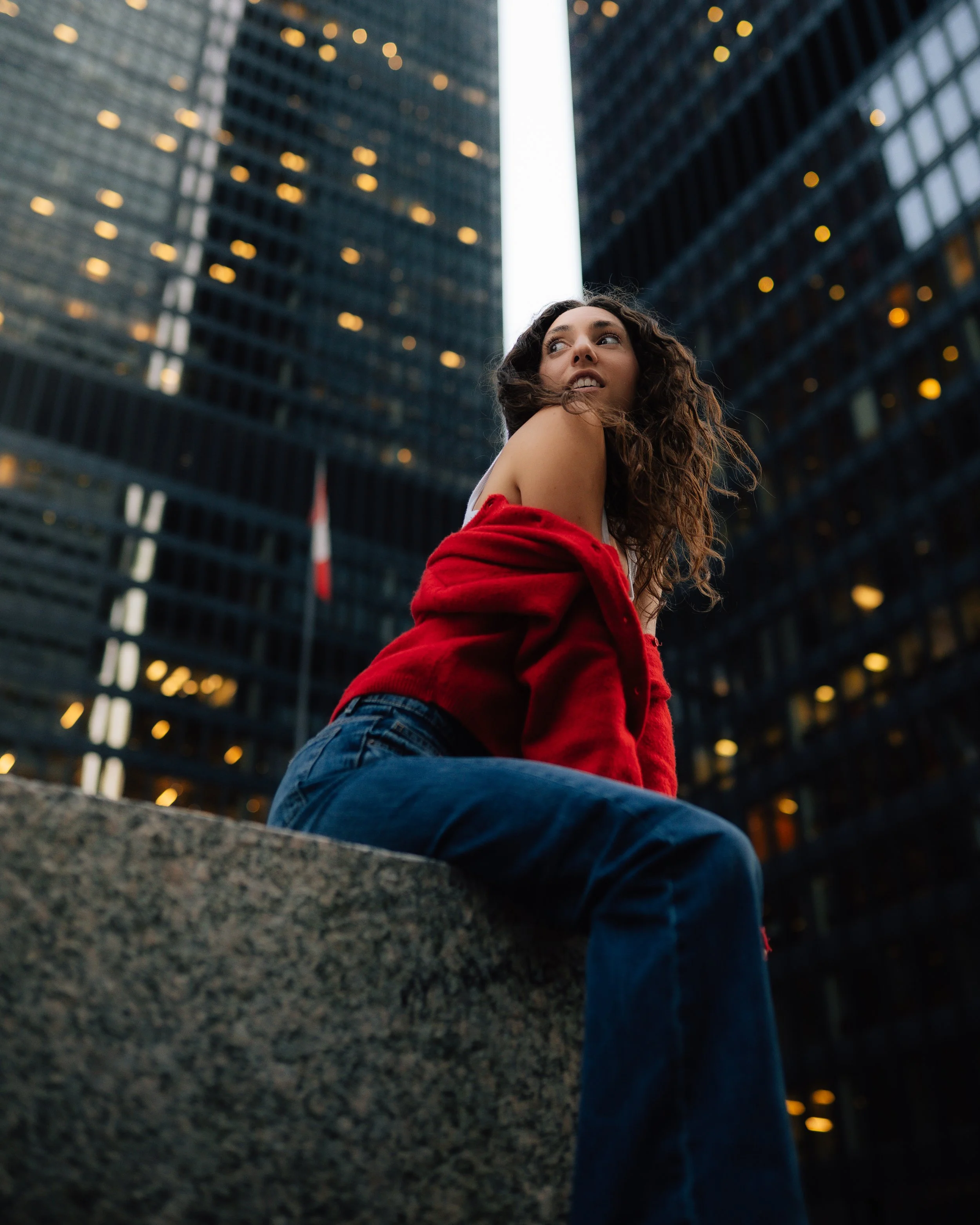 A young woman with curly hair sitting on a stone ledge in front of tall glass skyscrapers at dusk, wearing a red jacket off her shoulders and blue jeans, looking to the side.