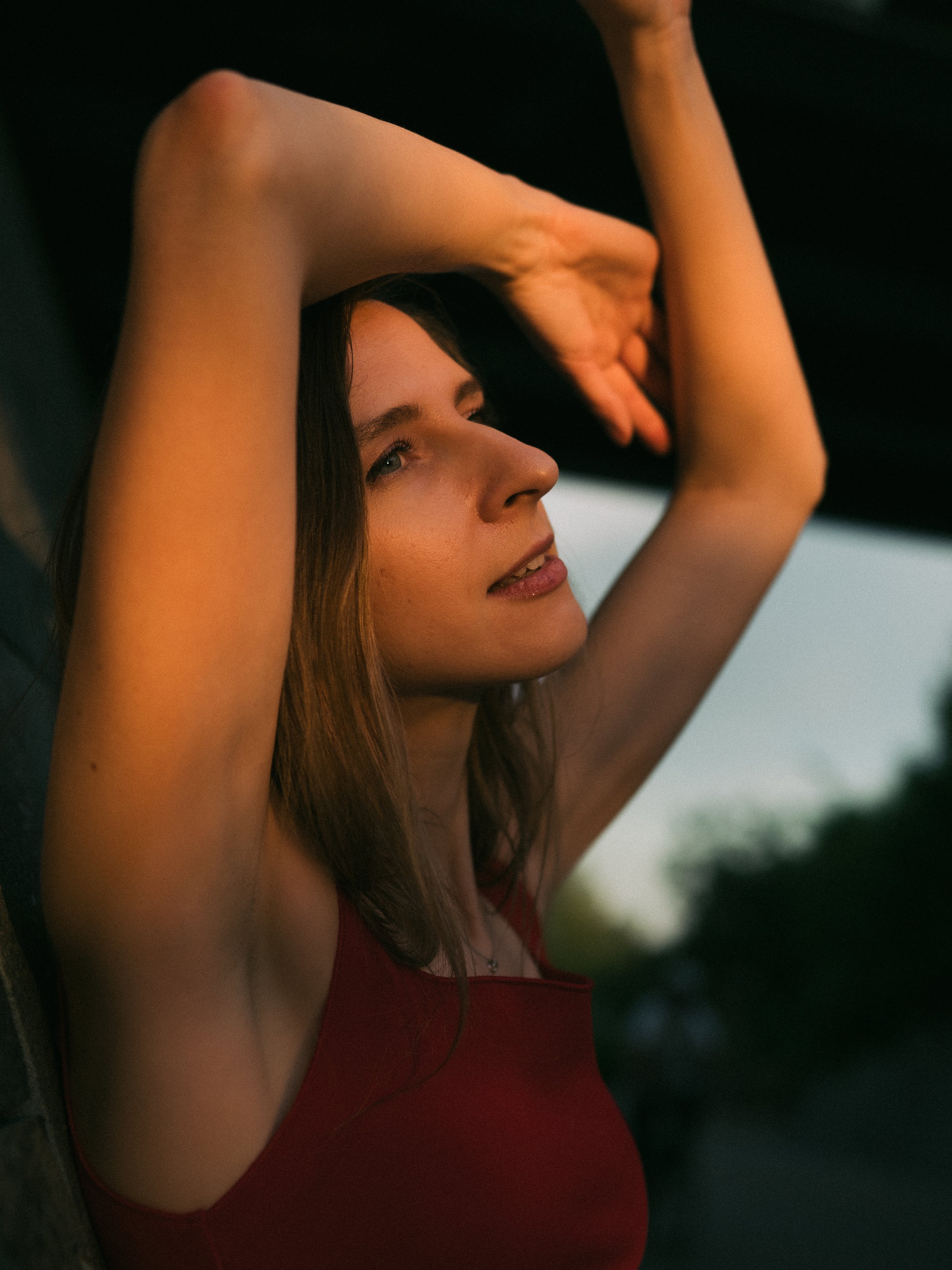 A woman with long brown hair and wearing a red top, standing outdoors at sunset, with her arms raised above her head, leaning against a dark surface.