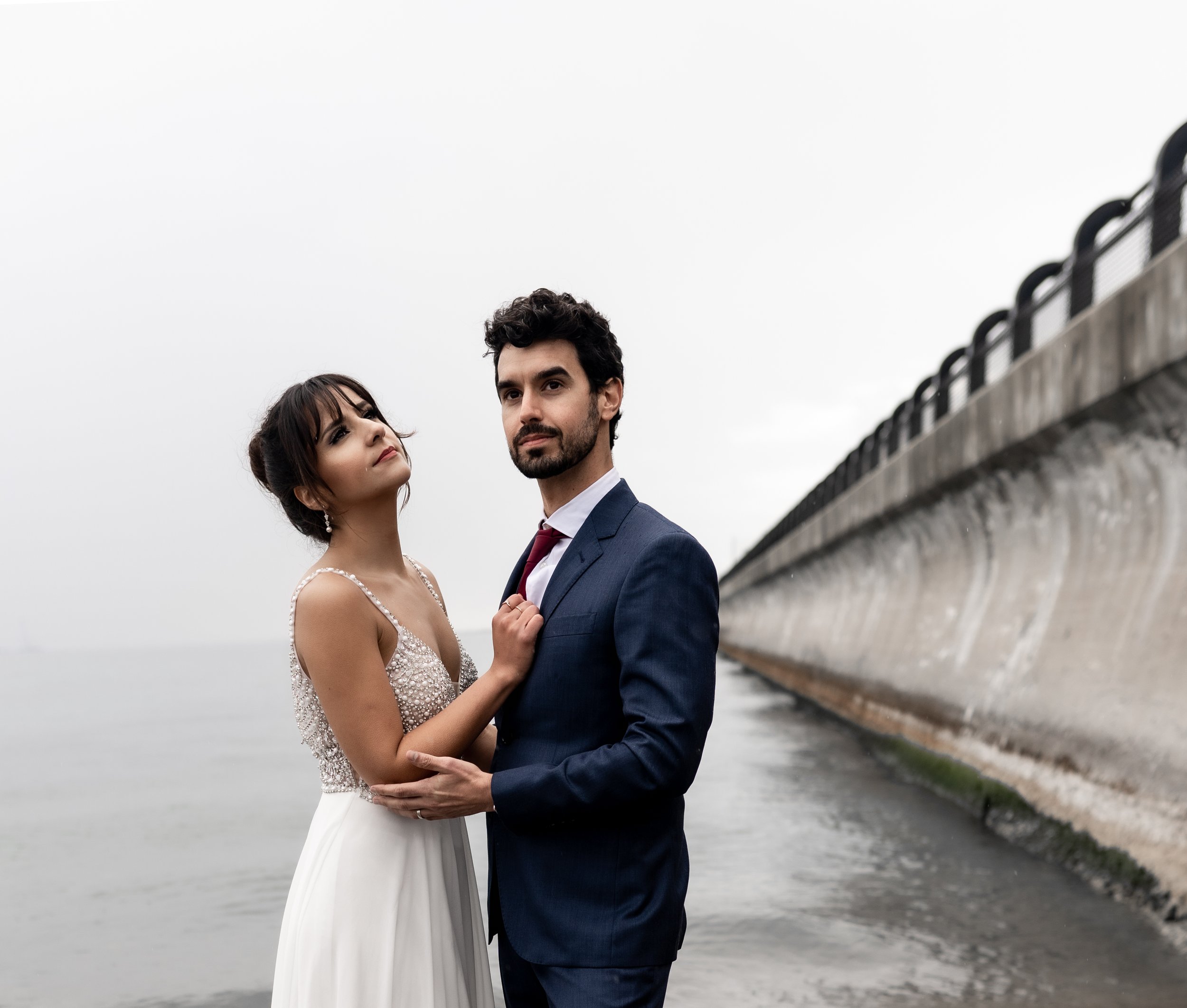 A couple standing by a waterfront, with the woman in a white dress and the man in a blue suit. The background shows a concrete wall and overcast sky.