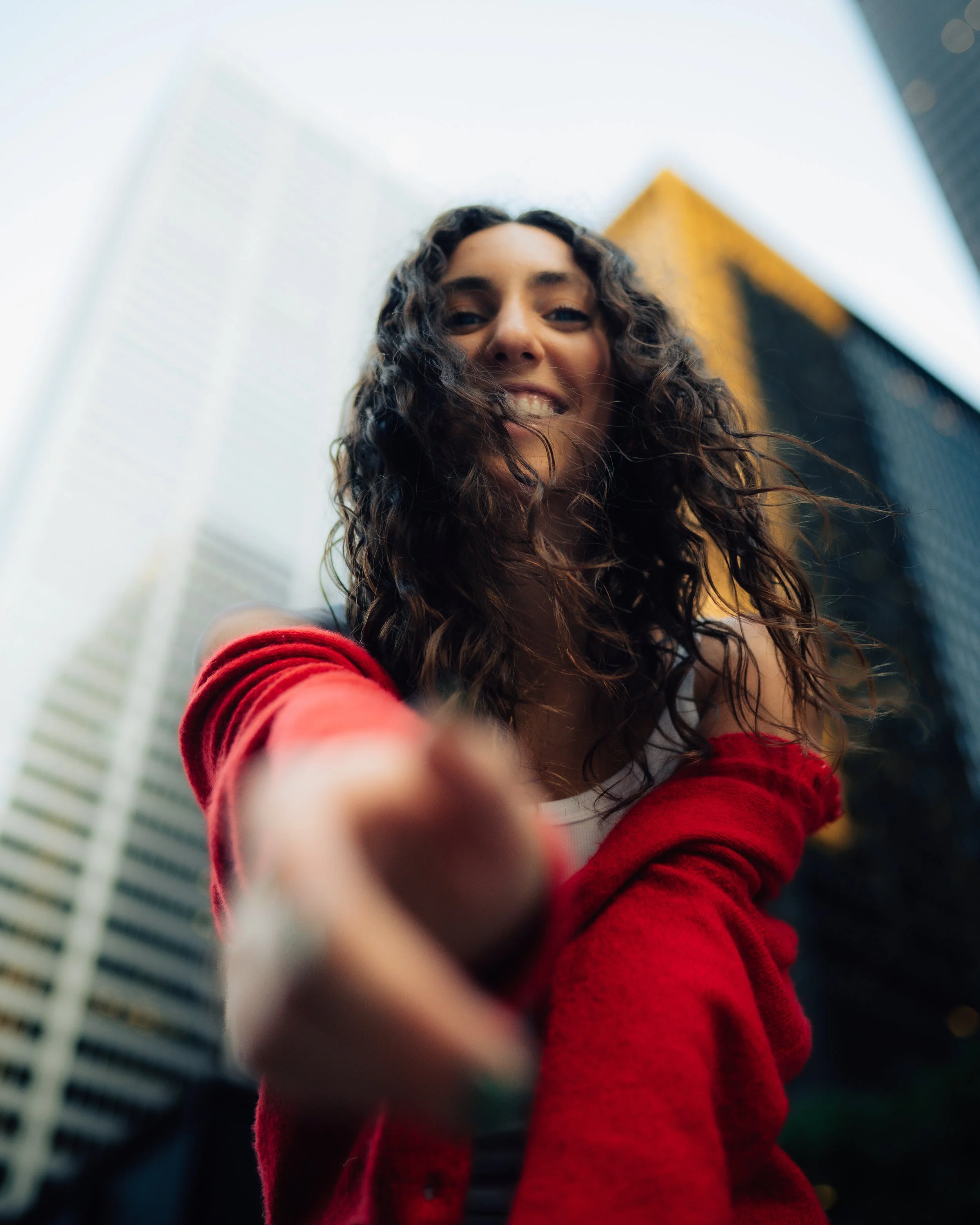 A young woman with curly hair wearing a red jacket, smiling and reaching toward the camera as she stands outdoors in a city with tall buildings in the background.