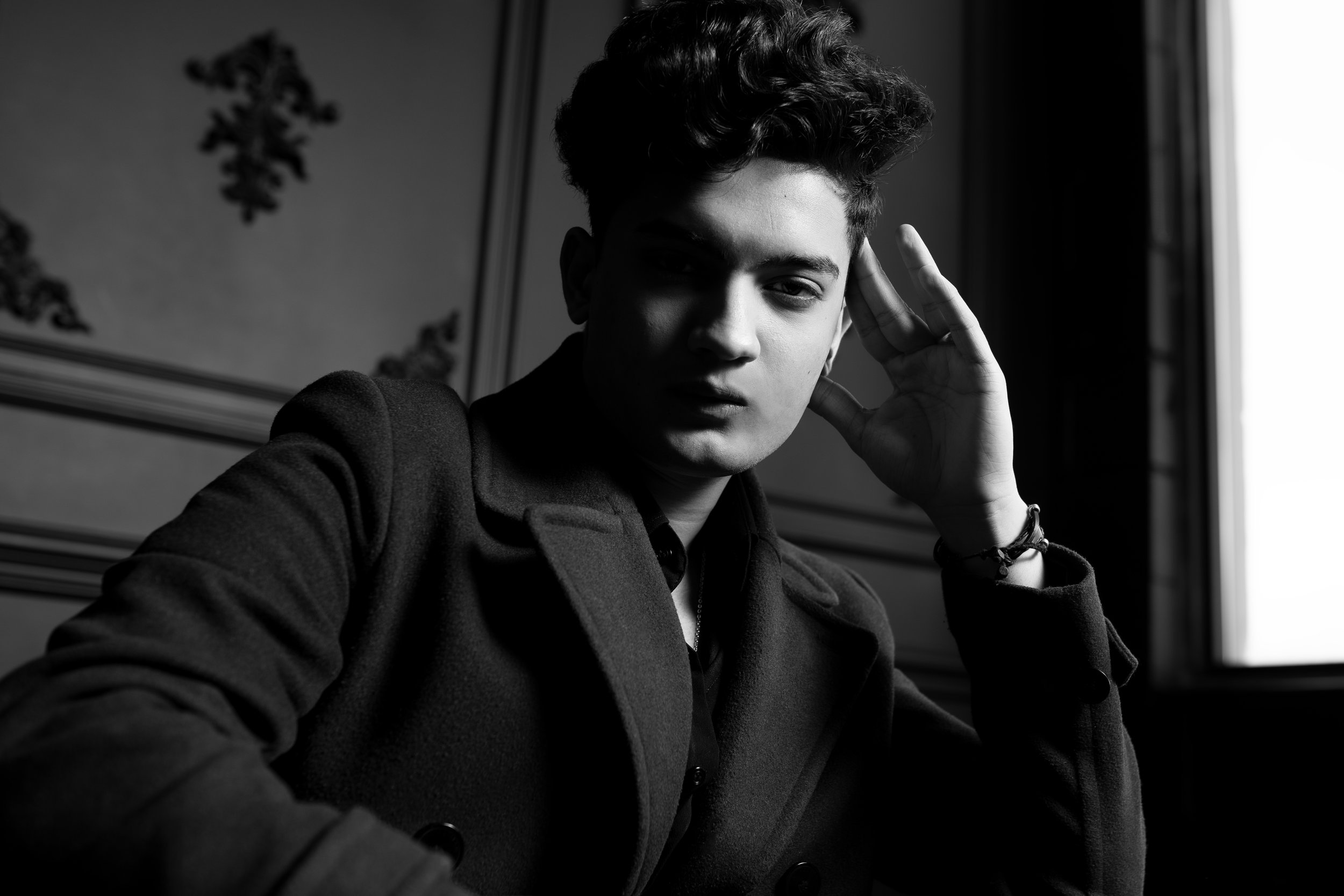 Black and white portrait of a young man in a dark coat, seated indoors, posing with one hand near his face.