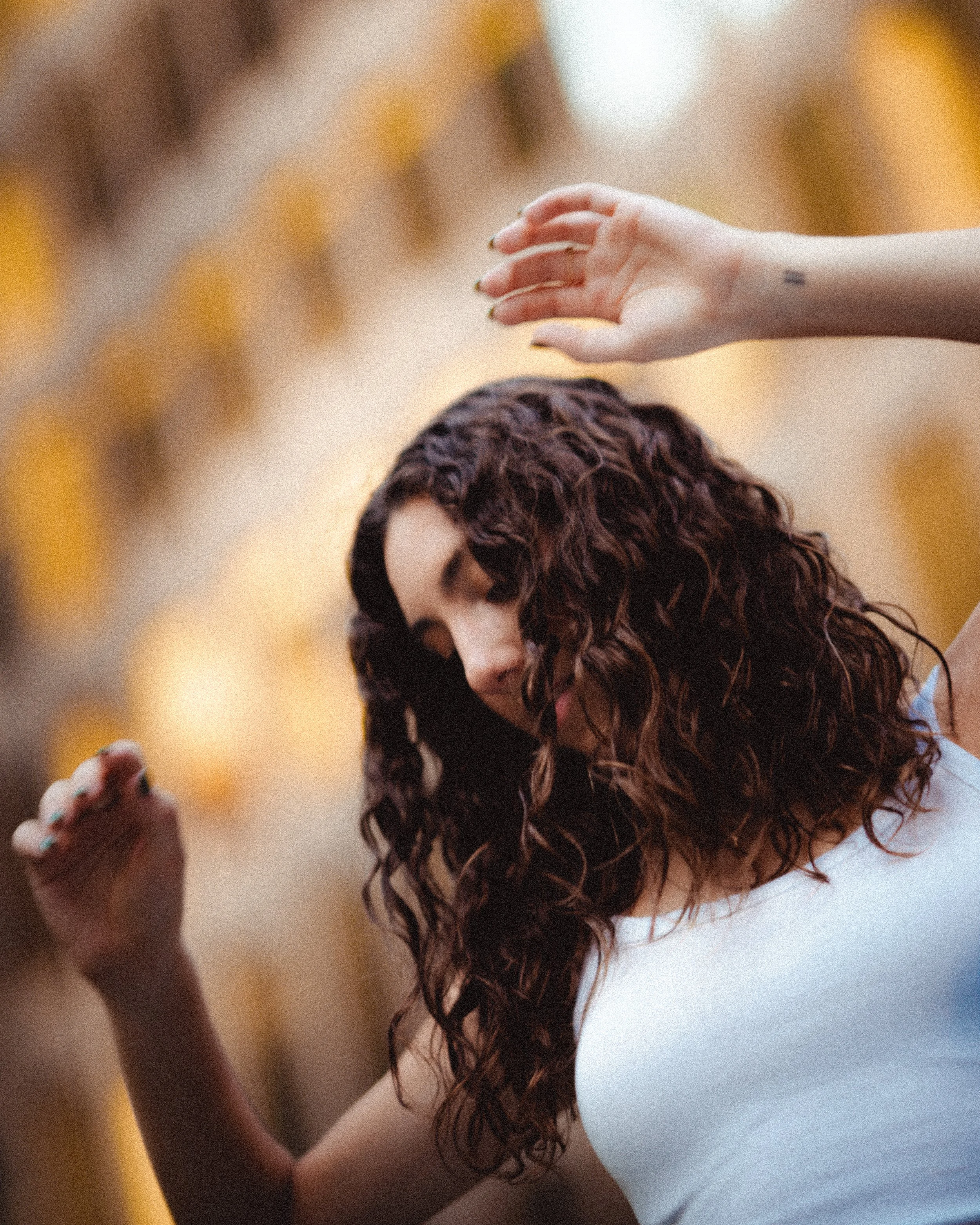 A woman with curly hair is dancing outdoors with a blurred, warm-colored background.