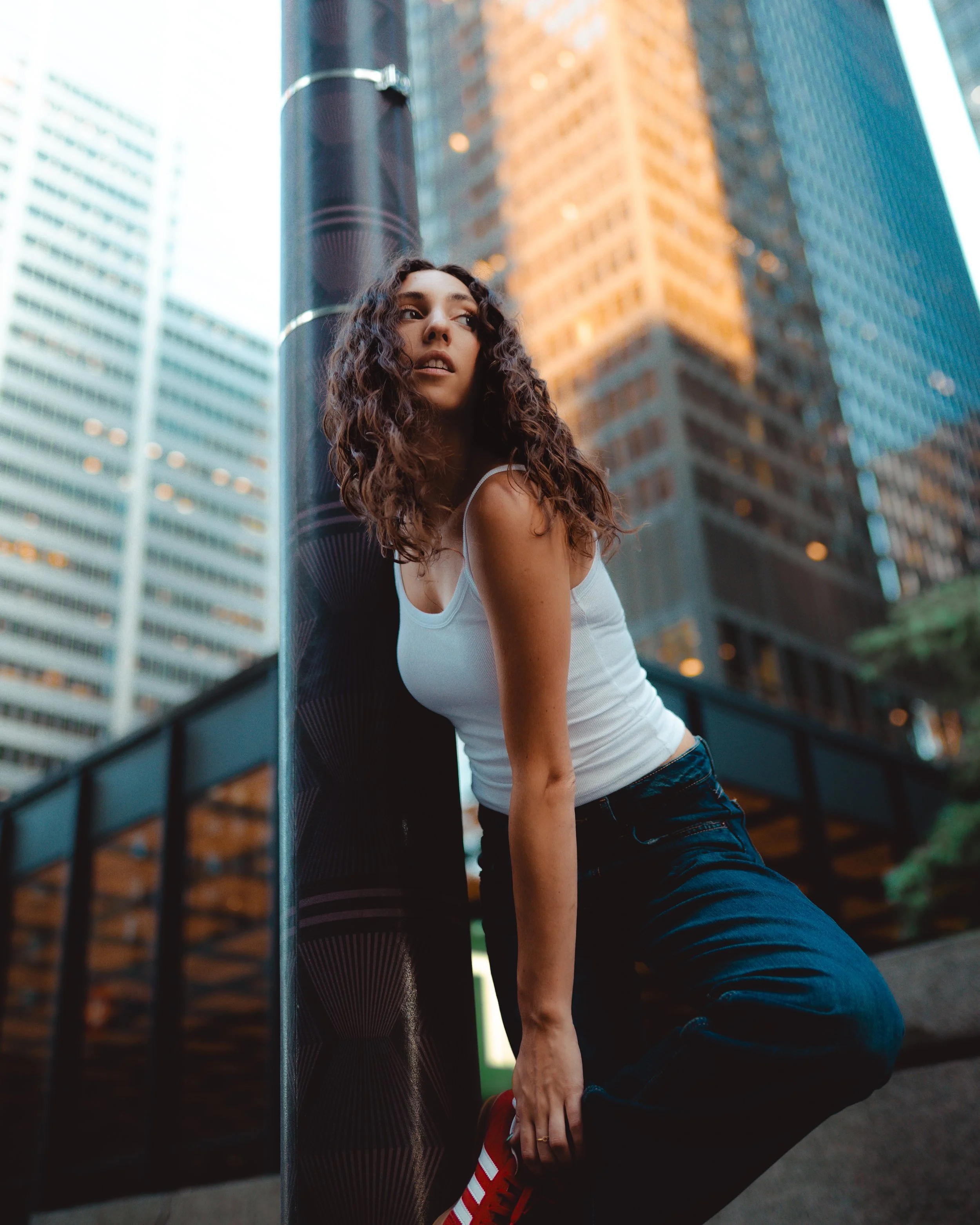 A young woman with curly brown hair wearing a white tank top and dark jeans, leaning against a black pole in an urban cityscape with tall buildings in the background.
