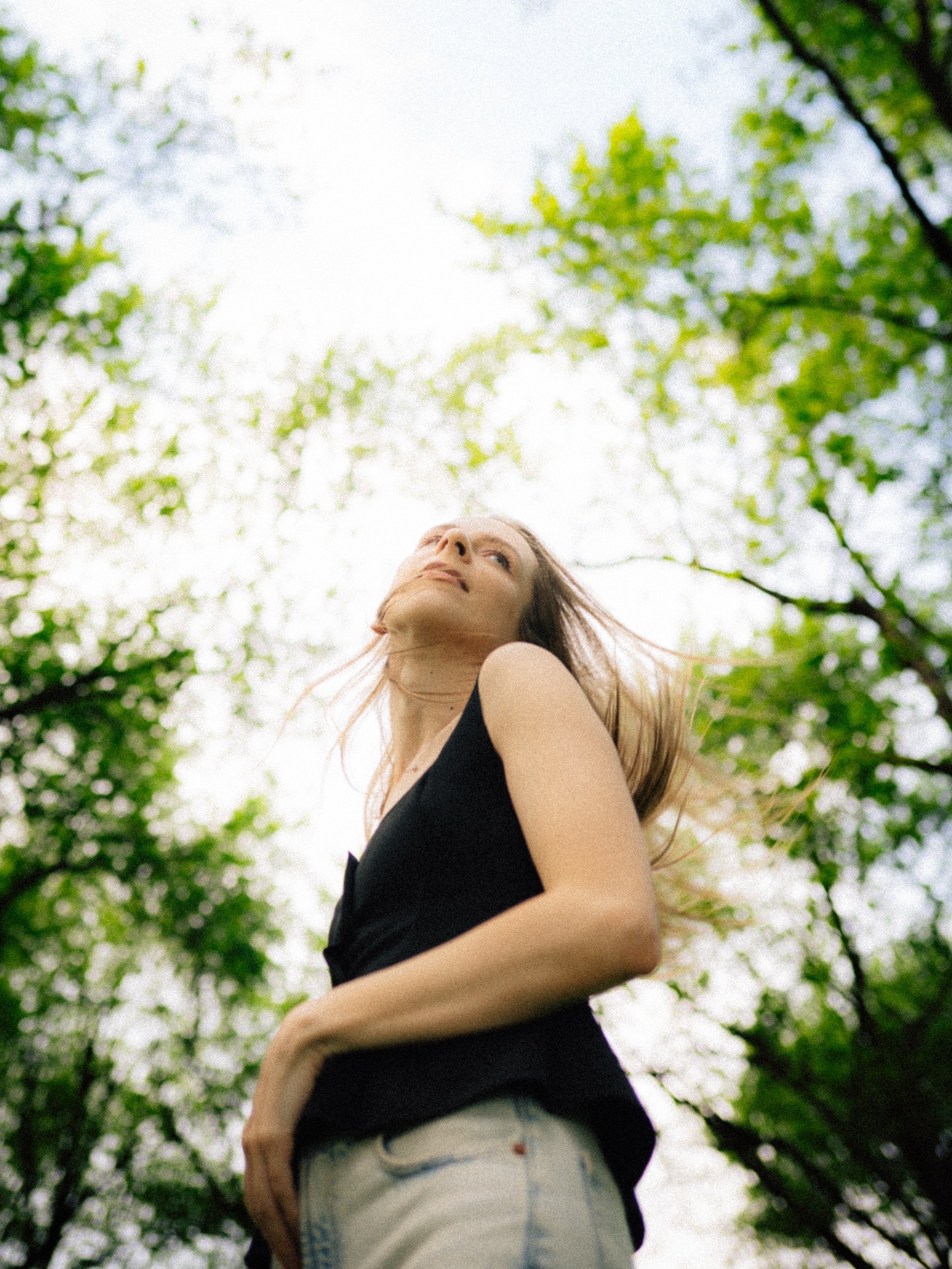 A woman with light skin and red hair standing outdoors amidst green trees, looking upwards, wearing a black sleeveless top and light-colored jeans.