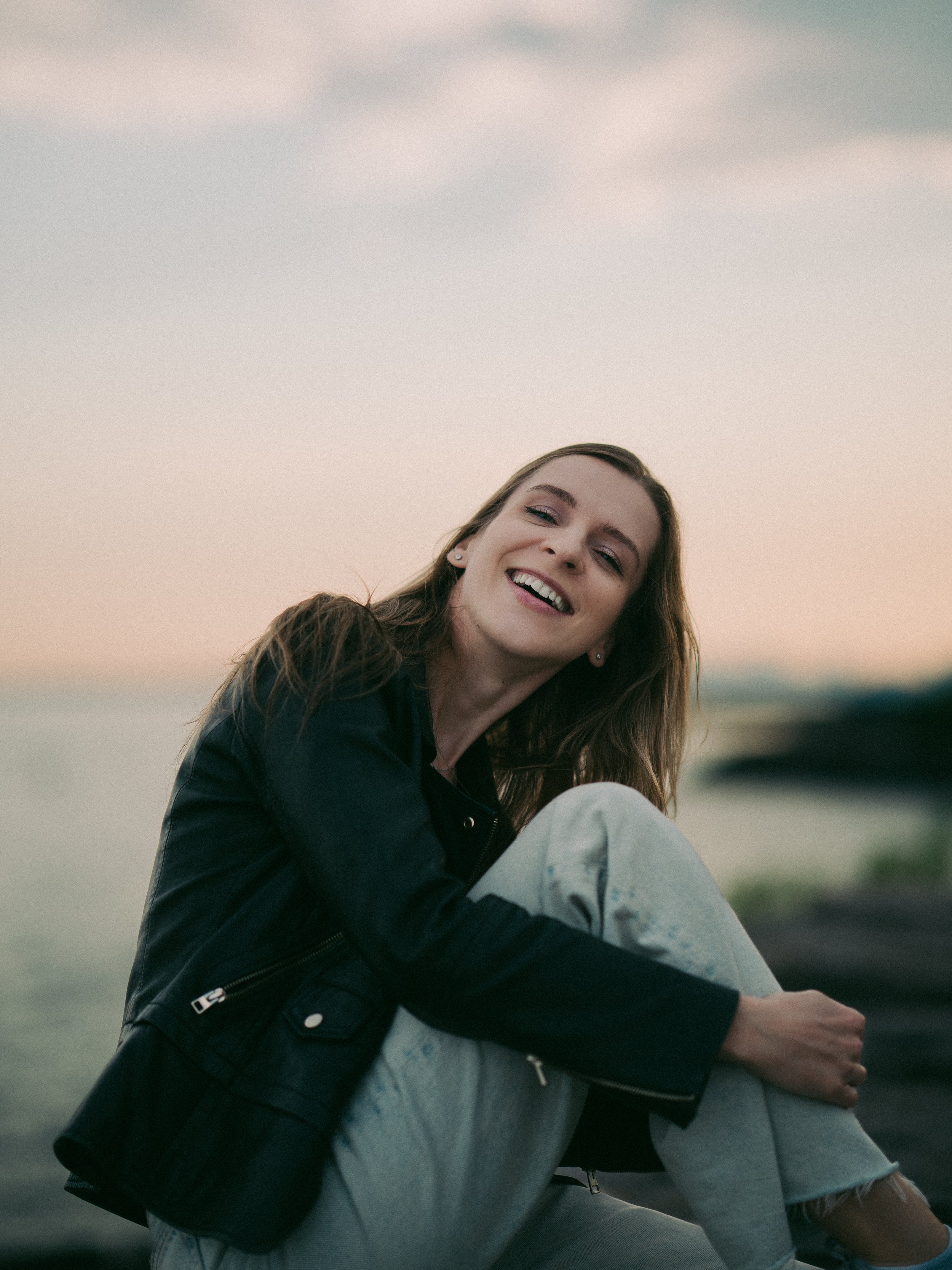 A smiling woman with long brown hair sitting outdoors near water at sunset, wearing a black leather jacket and light-colored pants.
