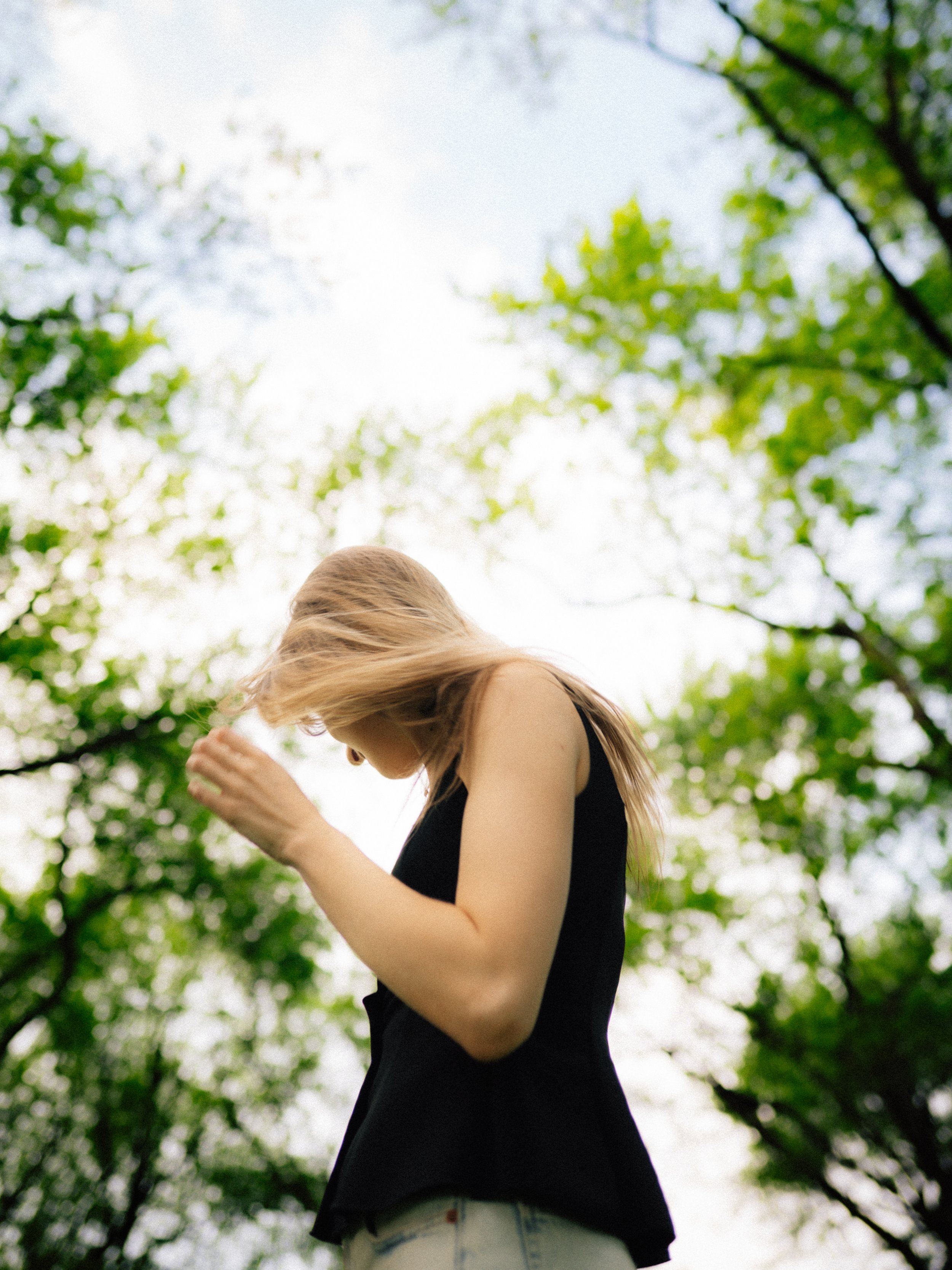 A woman with blonde hair wearing a black sleeveless top and light-colored jeans standing outdoors among green trees, with sunlight filtering through the leaves.