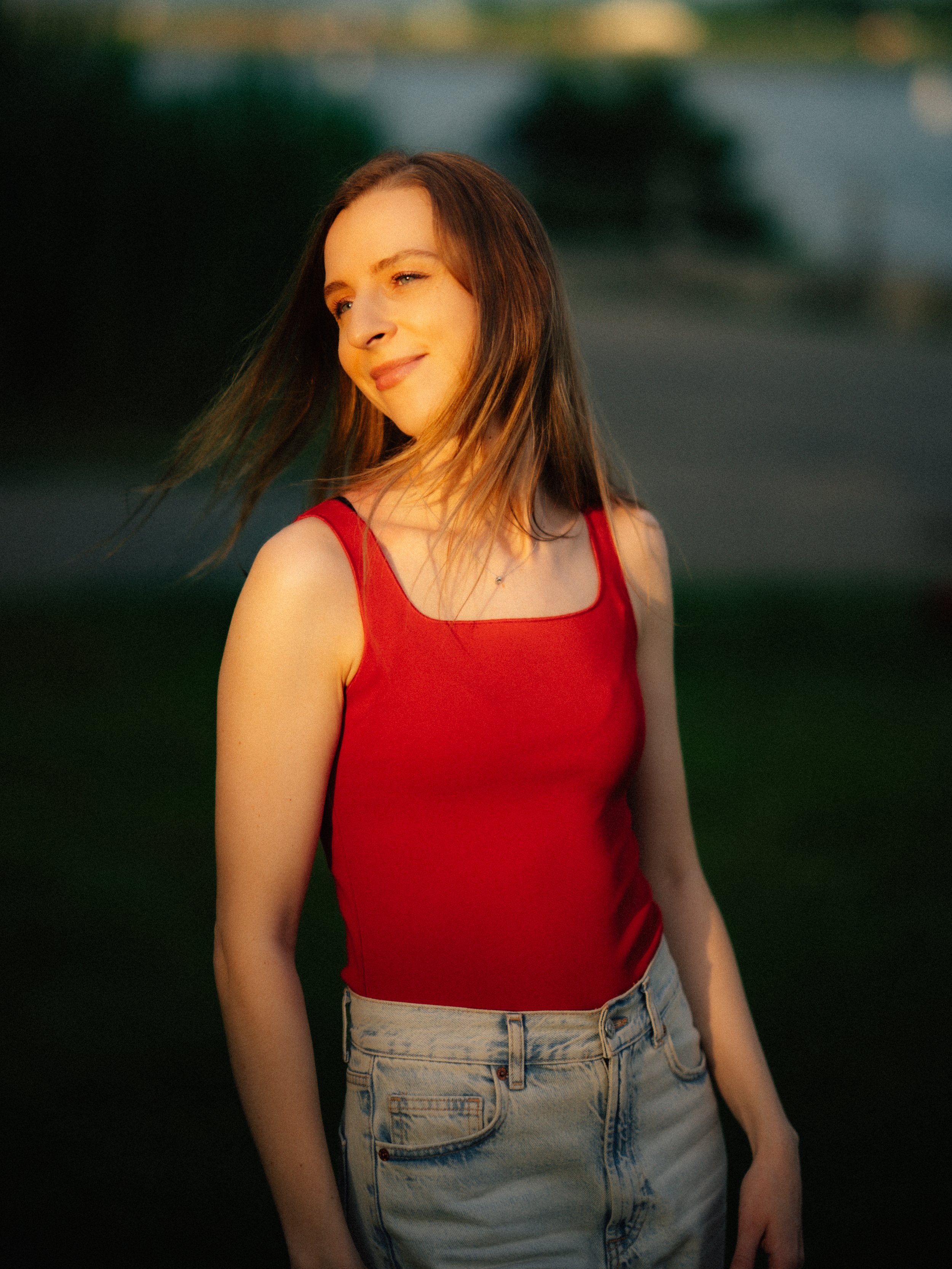 Young woman with brown hair wearing a red top and light denim jeans outdoors during sunset