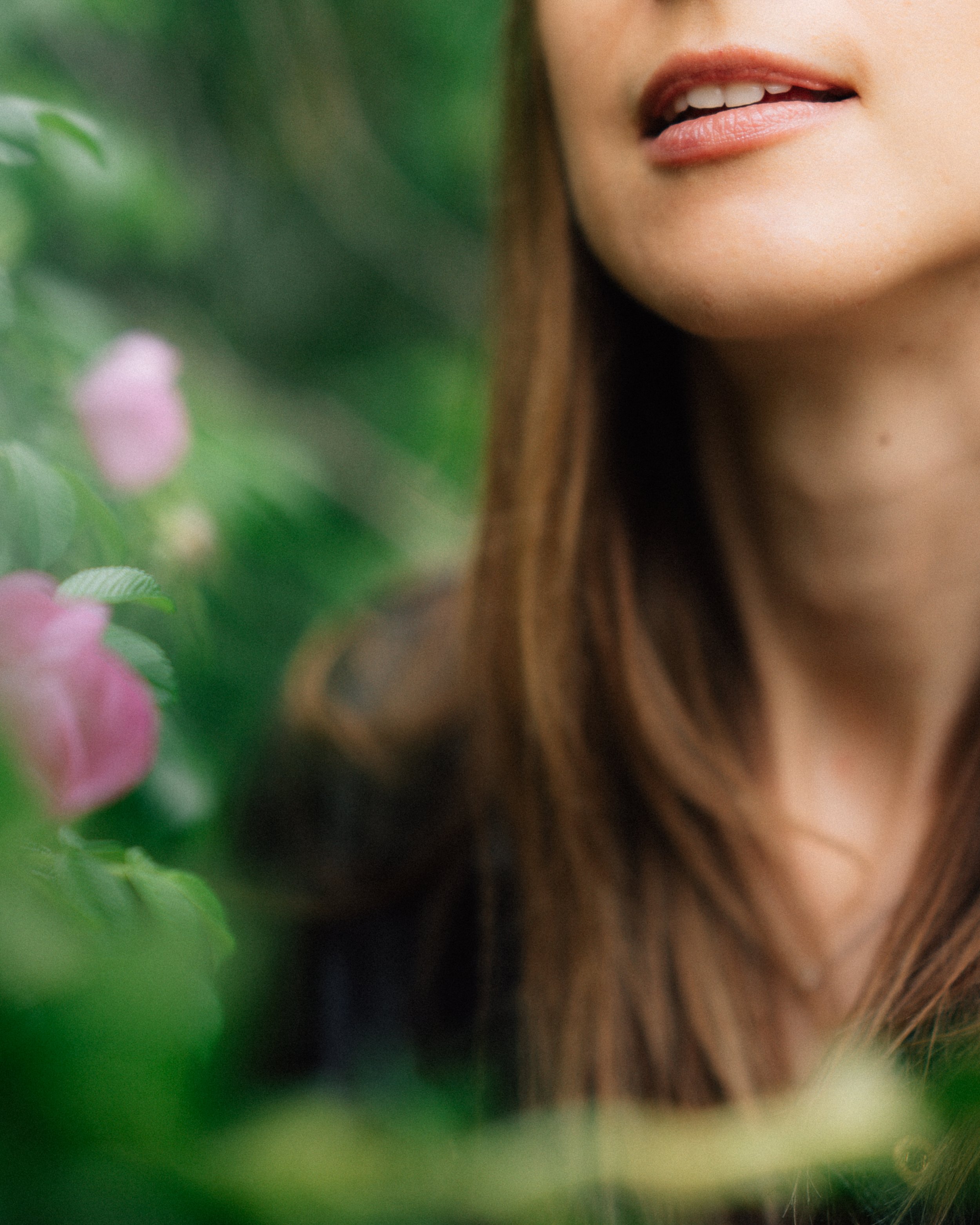 A close-up of a woman's face showing her lips and chin, with her long brown hair, surrounded by and partially obscured by green foliage and pink flowers.