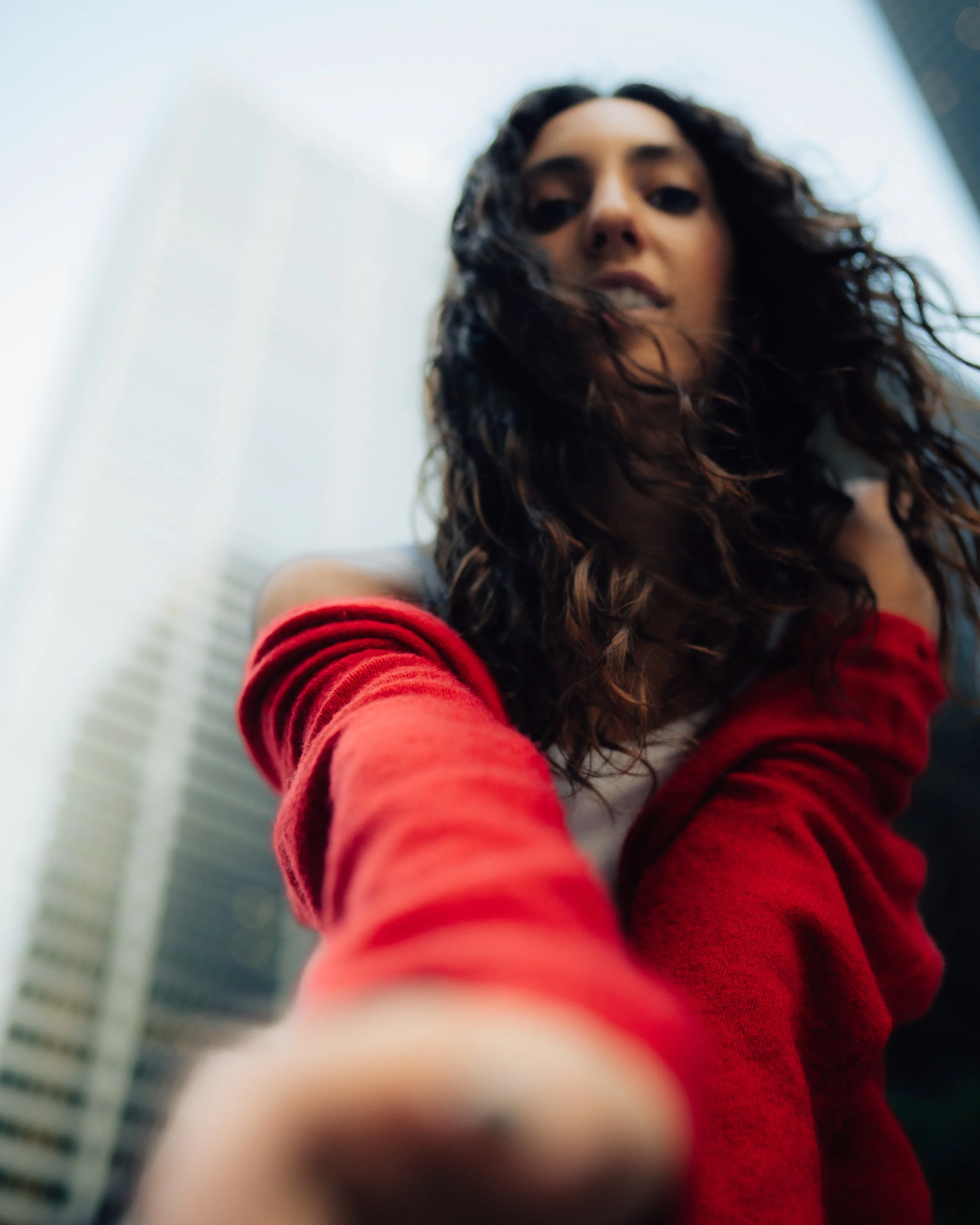 A woman with curly hair wearing a red jacket takes a low-angle selfie against a city skyline with tall buildings and a cloudy sky.