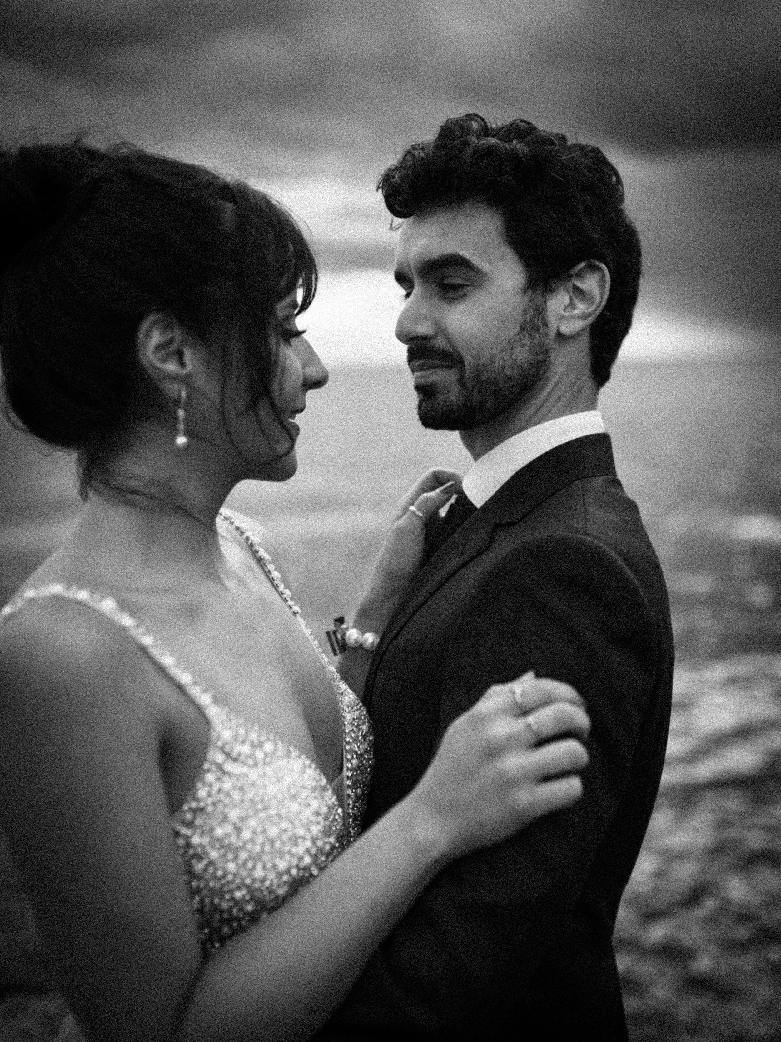 A black-and-white photo of a couple in formal attire, standing close and gazing at each other, with a body of water in the background.