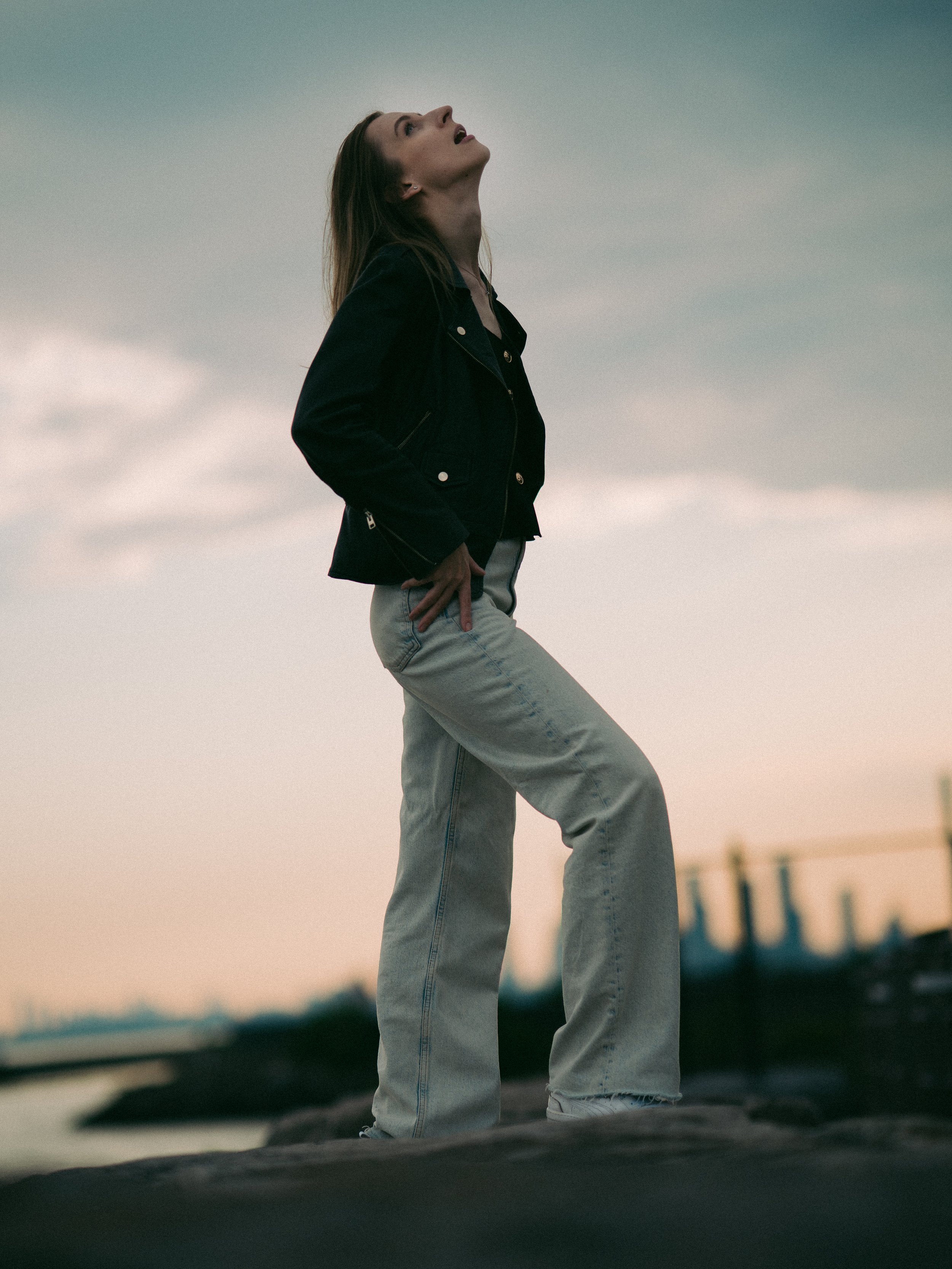 A woman wearing a black jacket and light-colored jeans standing outdoors during sunset, looking upwards with her hands on her hips.