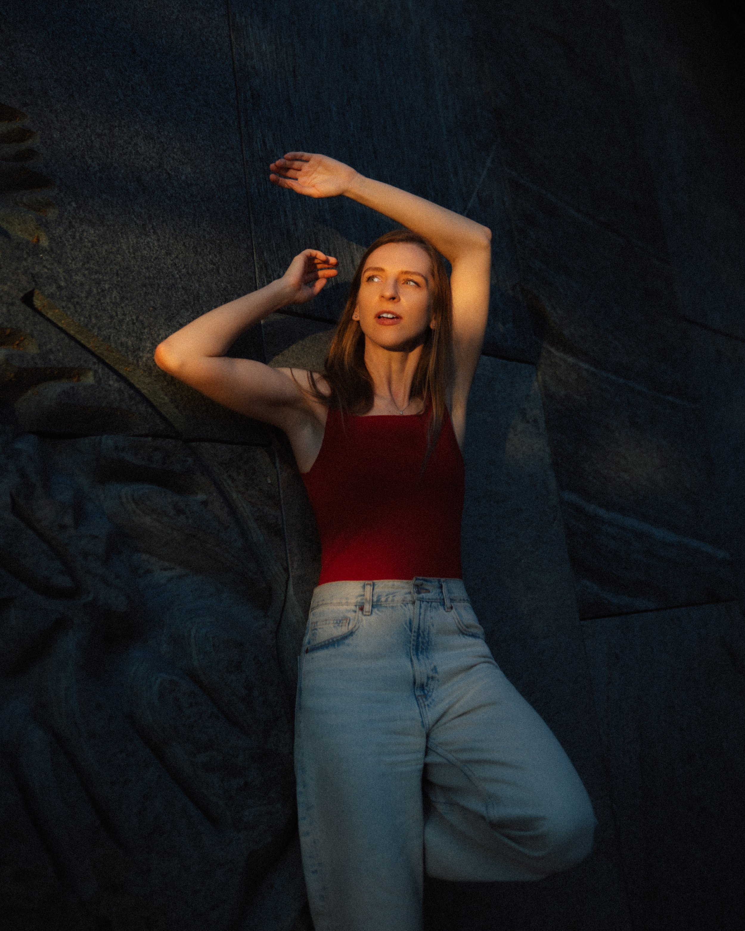 A woman with long brown hair wearing a red sleeveless top and light blue jeans is lying on a dark stone surface, with her arms raised above her head, illuminated by warm lighting.