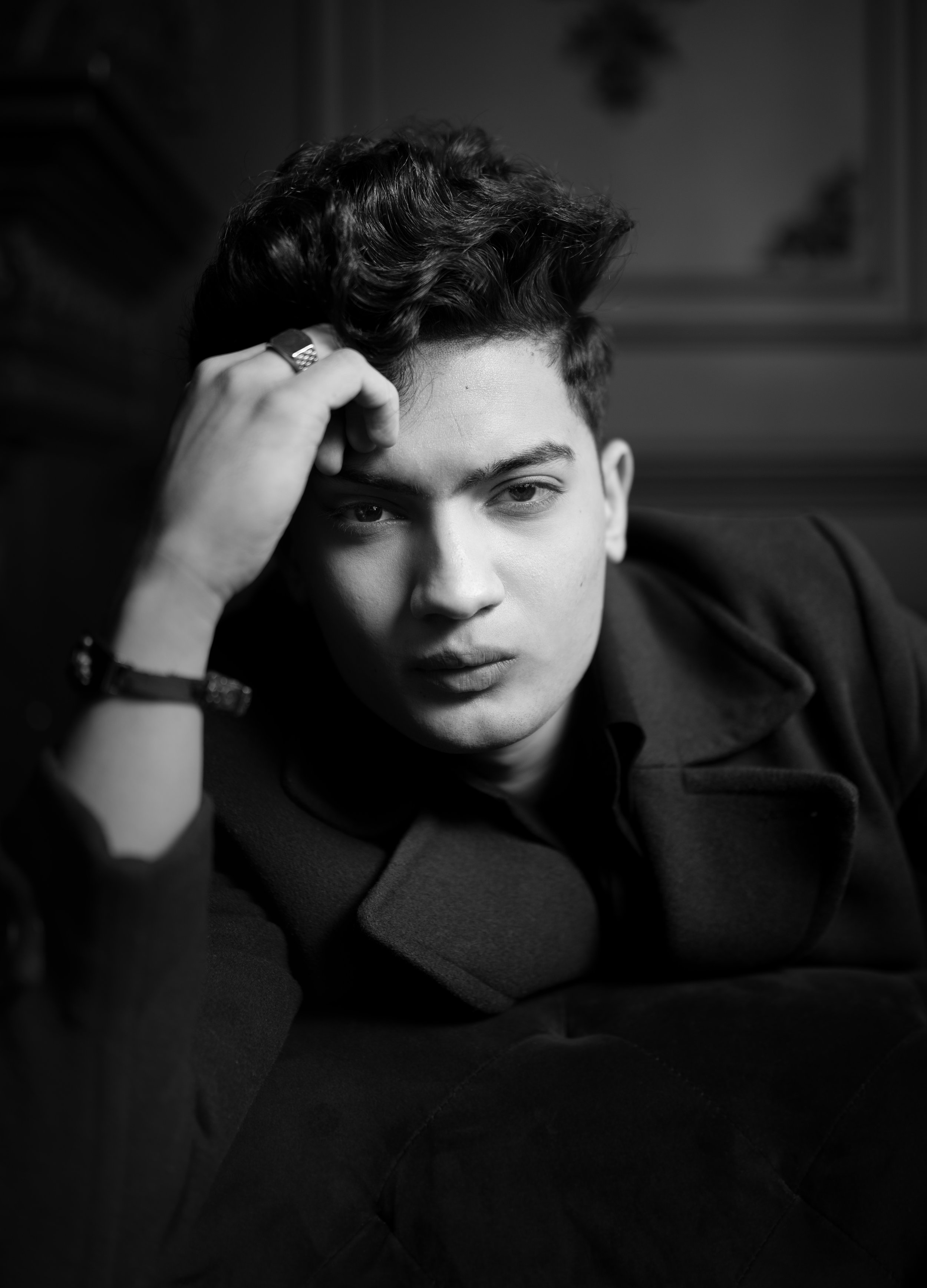 A black and white portrait of a young man with styled hair, looking directly at the camera, resting his head on his hand, wearing a dark coat or jacket.