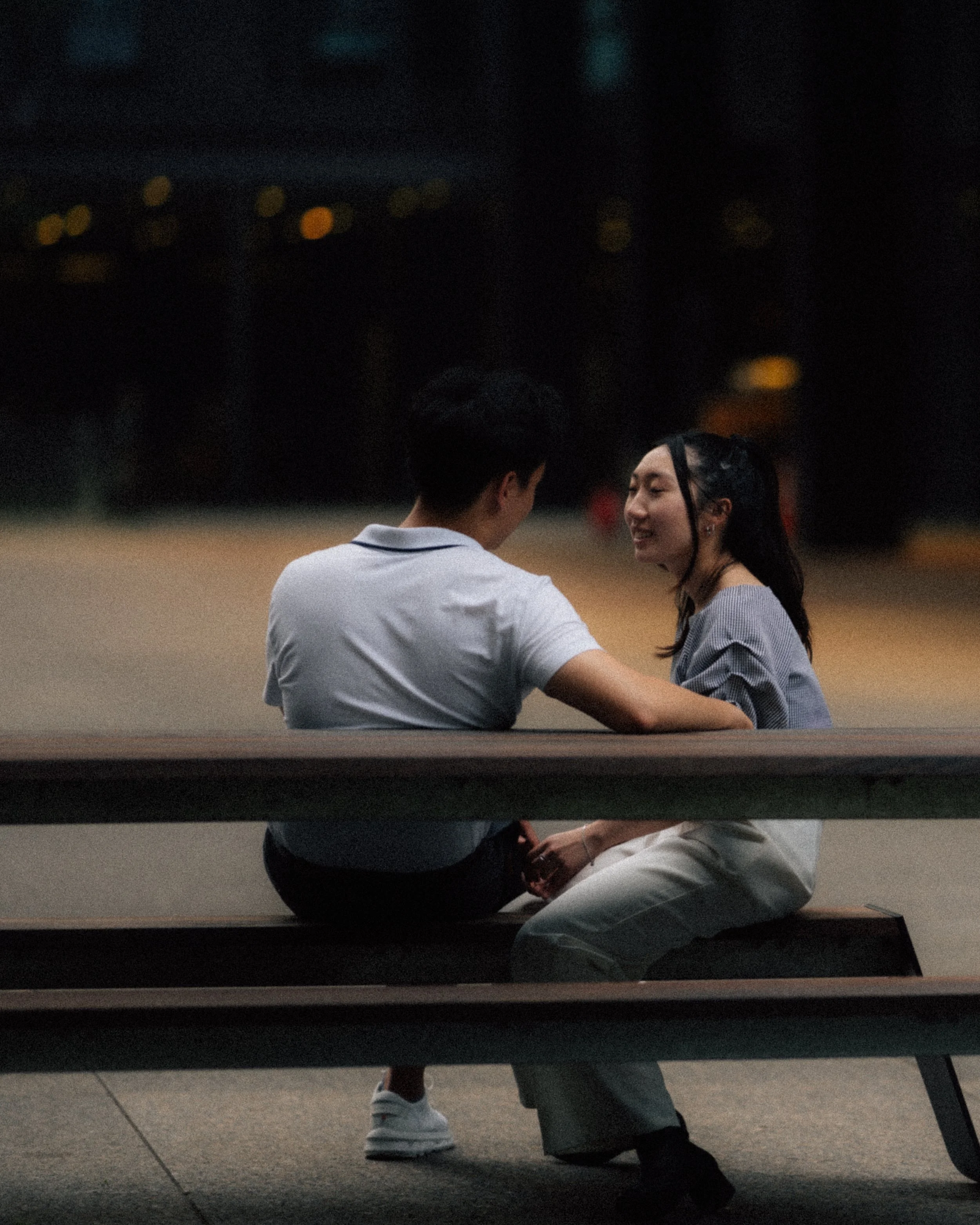 A young man and woman sitting on a park bench at night, facing each other and sharing a moment of connection.