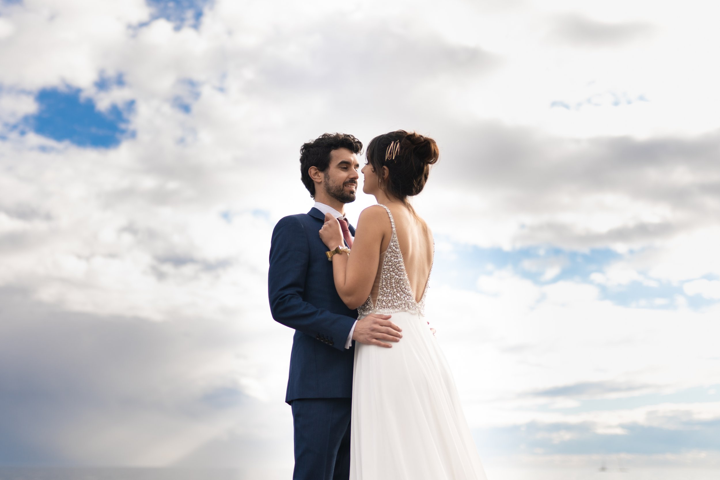Bride and groom embracing outdoors under cloudy sky.