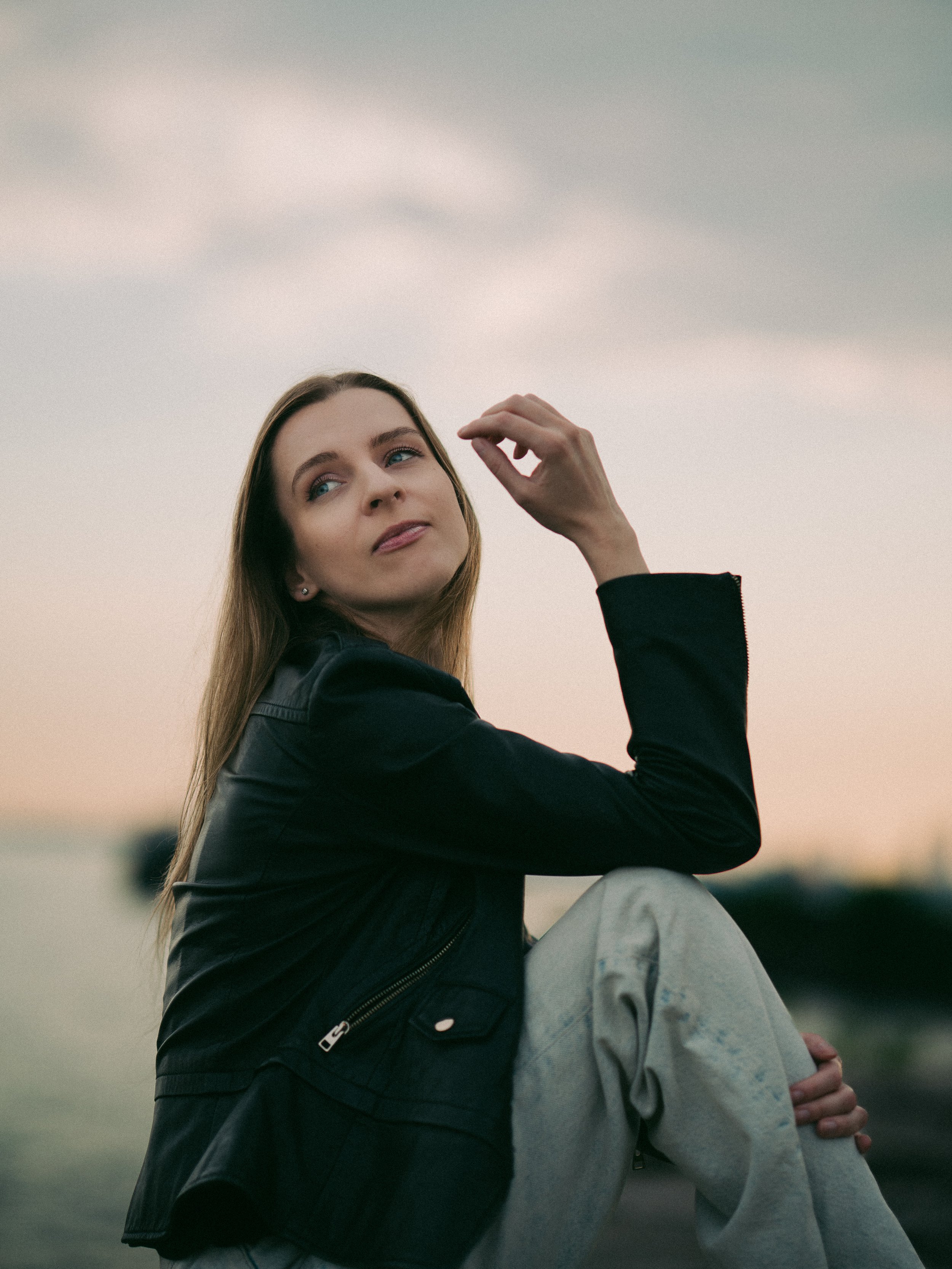A young woman with long, light brown hair sits outdoors at sunset, wearing a black leather jacket and light-colored pants, gazing thoughtfully into the distance.