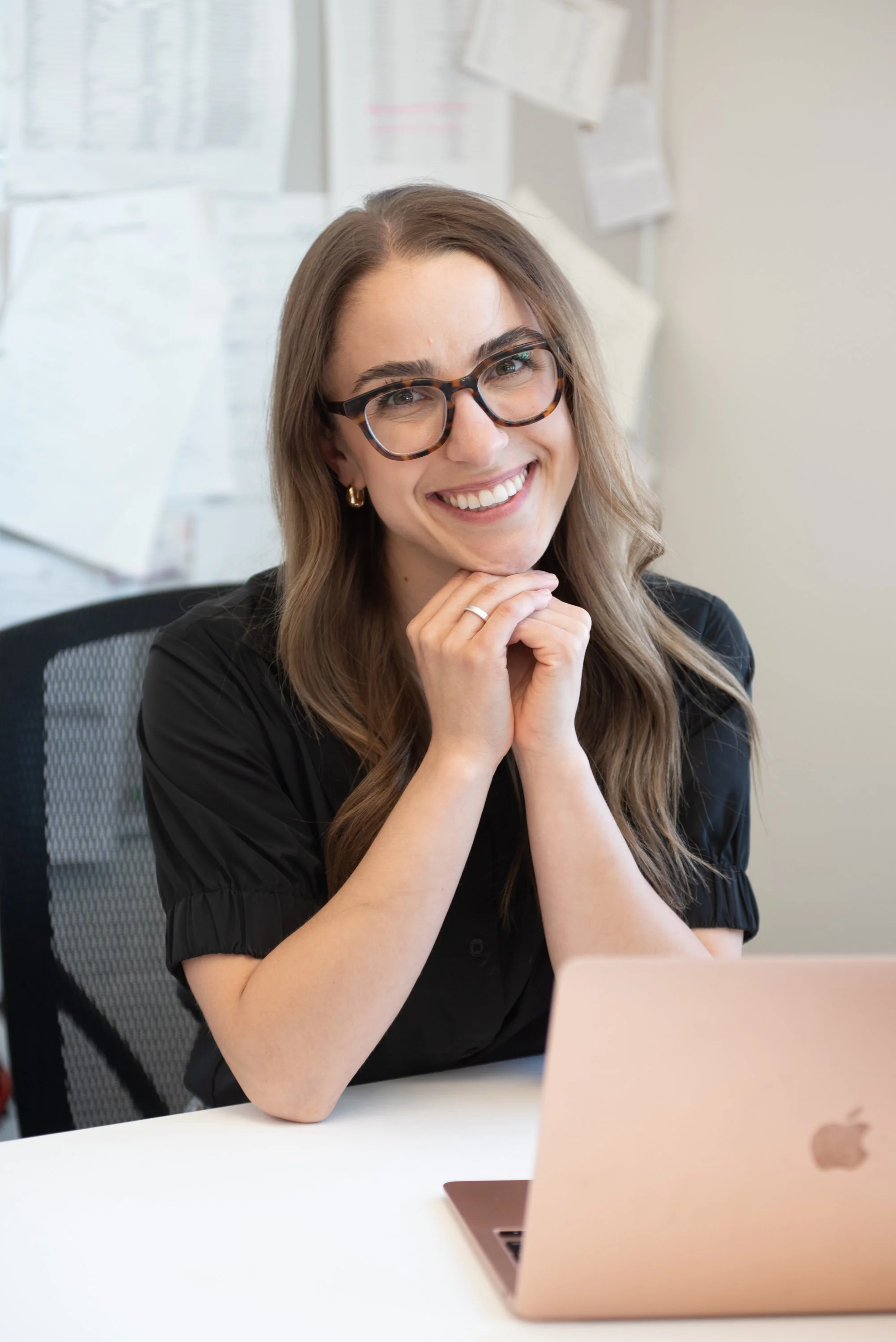 Smiling woman with glasses sitting at a desk with a laptop, hands clasped under her chin, and papers on the wall in the background.