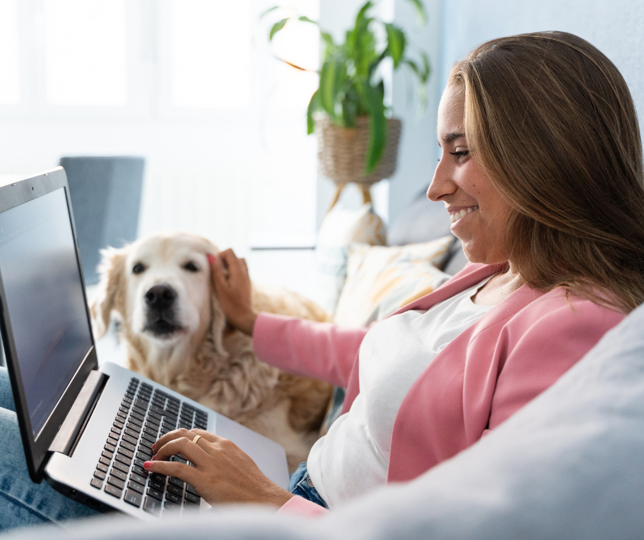 A woman smiling while sitting on a couch using a laptop and petting a golden retriever dog.