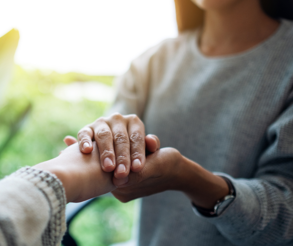 Person gently holding a child's hand outdoors, with greenery and sunlight in the background.