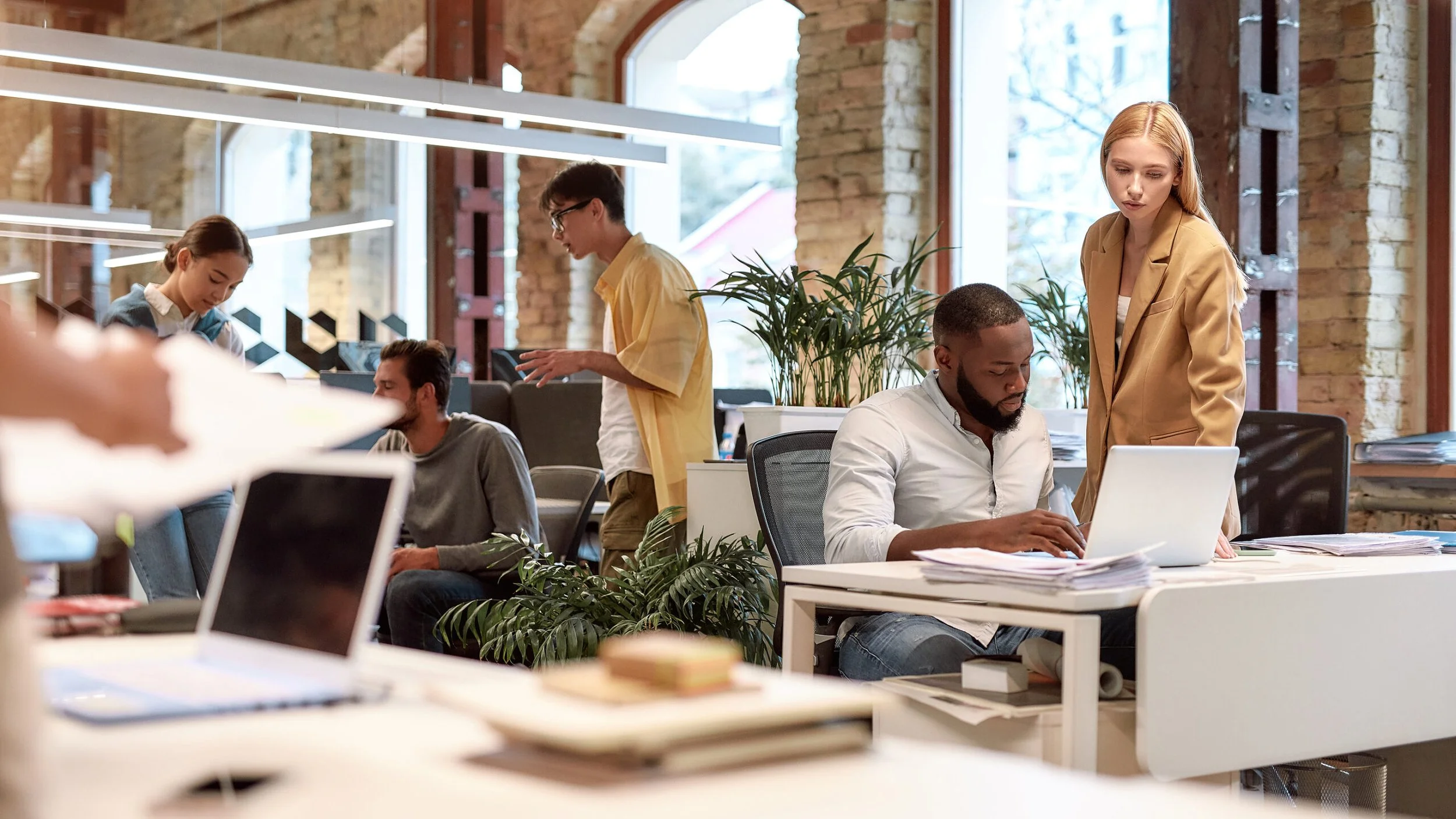 Modern open-plan office with diverse people working, laptops on desks, brick walls, and large windows.
