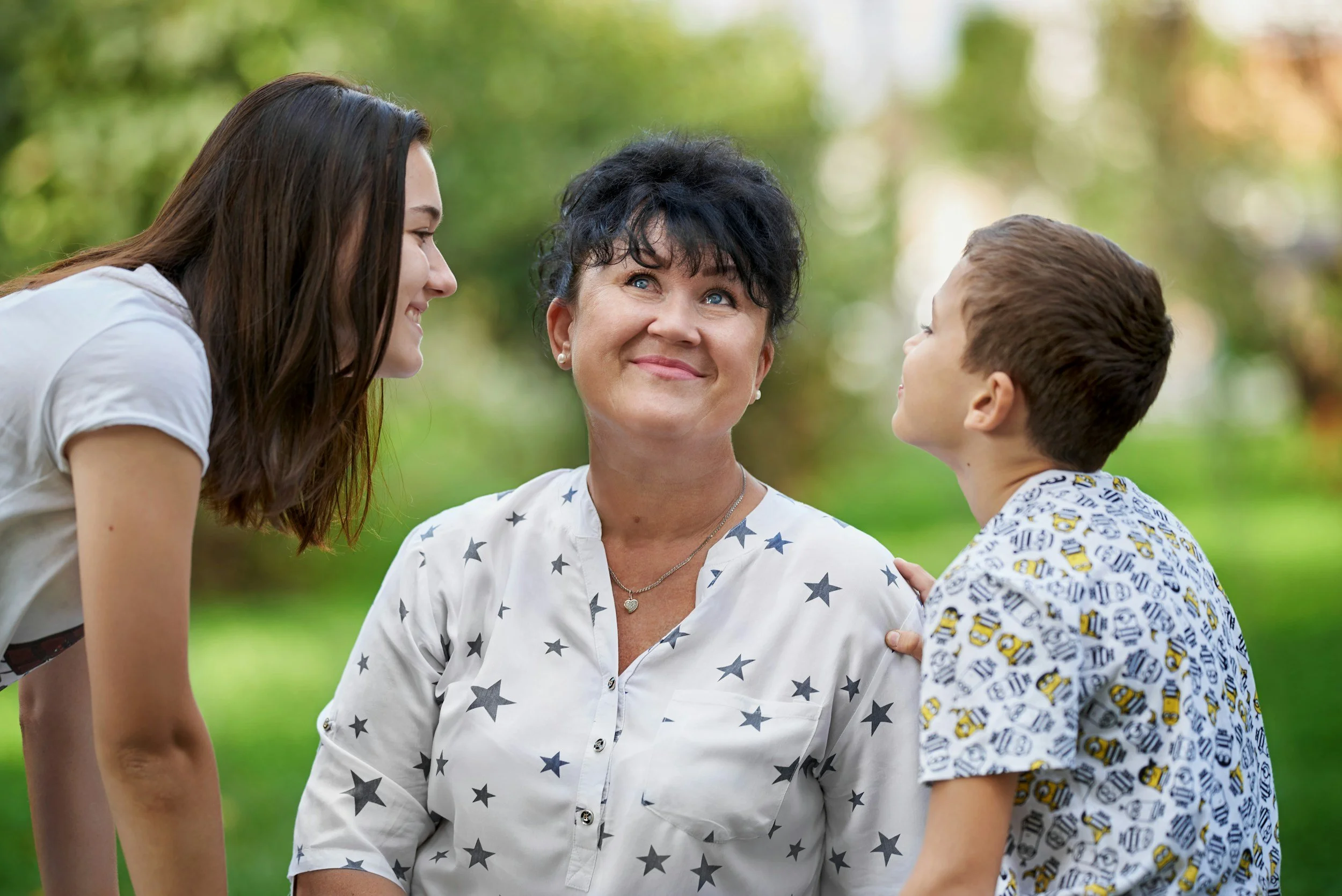 A woman in a star-patterned shirt smiling, surrounded by a girl on her left and a boy on her right, in an outdoor setting.