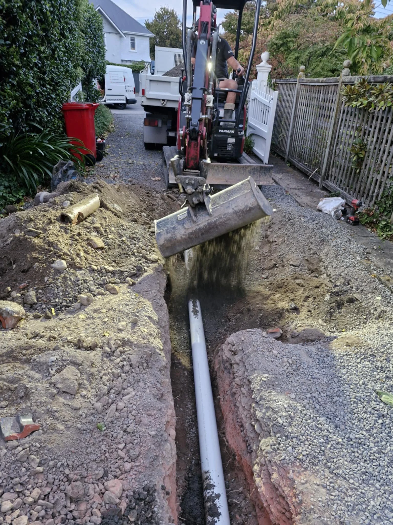 Mini excavator filling a trench with soil over a newly installed pipe in a driveway.