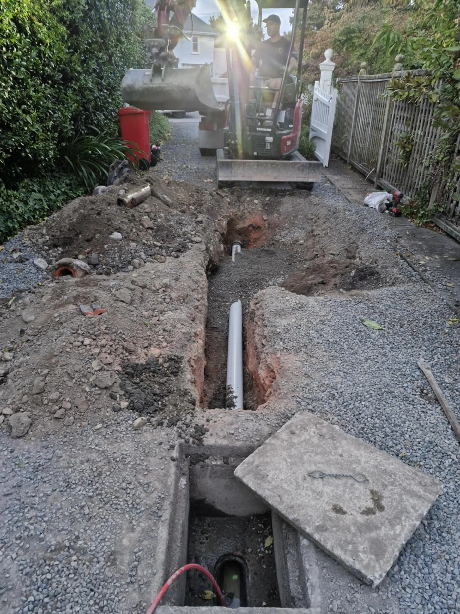 Excavator working on trench in driveway for drainage installation, with exposed pipe and gravel.