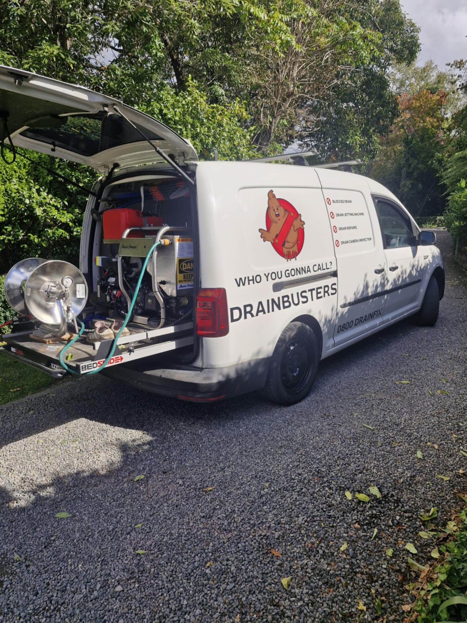 A white van with the back door open displays equipment inside. The side of the van features a logo and services offered, including drain unblocking and inspections, under the name 'Drainbusters.' The van is parked on a gravel driveway, surrounded by greenery.