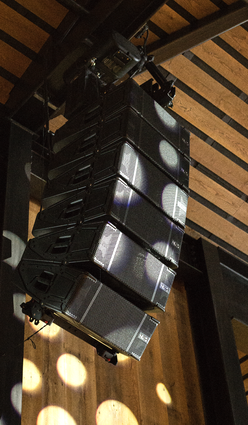 Multiple black speakers mounted on a wooden ceiling in a music or event venue.