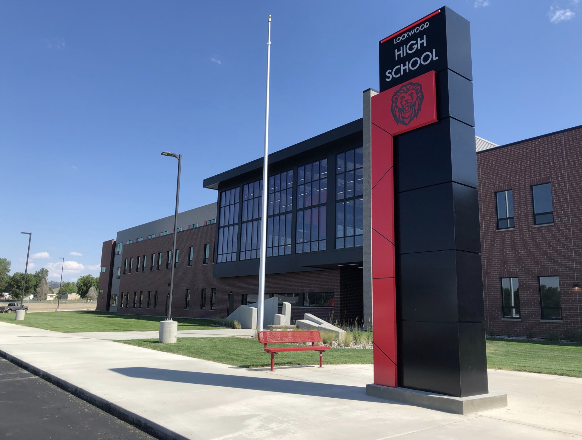Exterior view of Lockwood High School building with a large sign featuring the school's name and lion logo, a red bench, and a sidewalk under a clear blue sky.