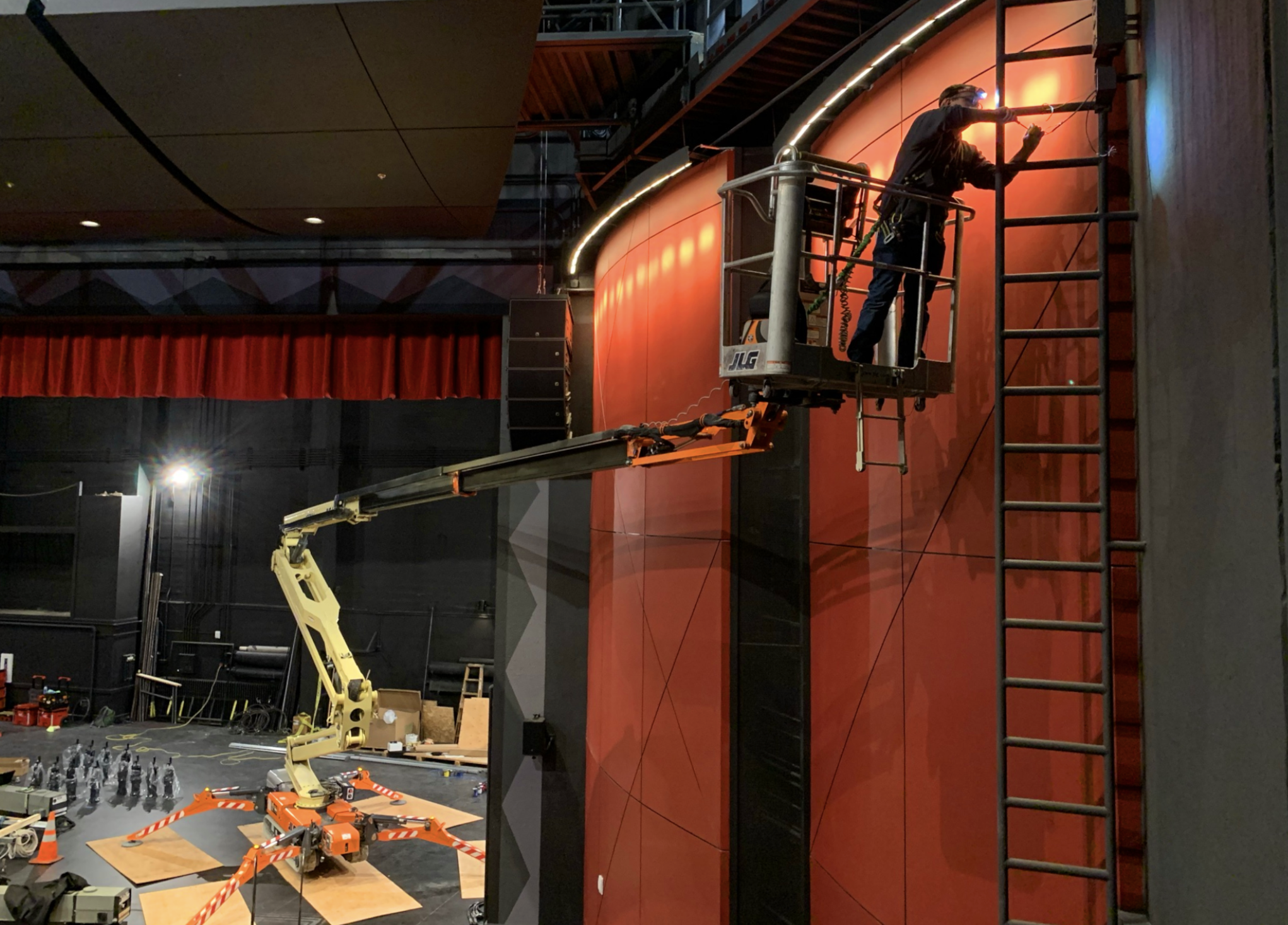A worker on a cherry picker high above the stage wall, painting or installing light fixtures. The stage area below has equipment, cones, and bottled water, inside a theater or auditorium.