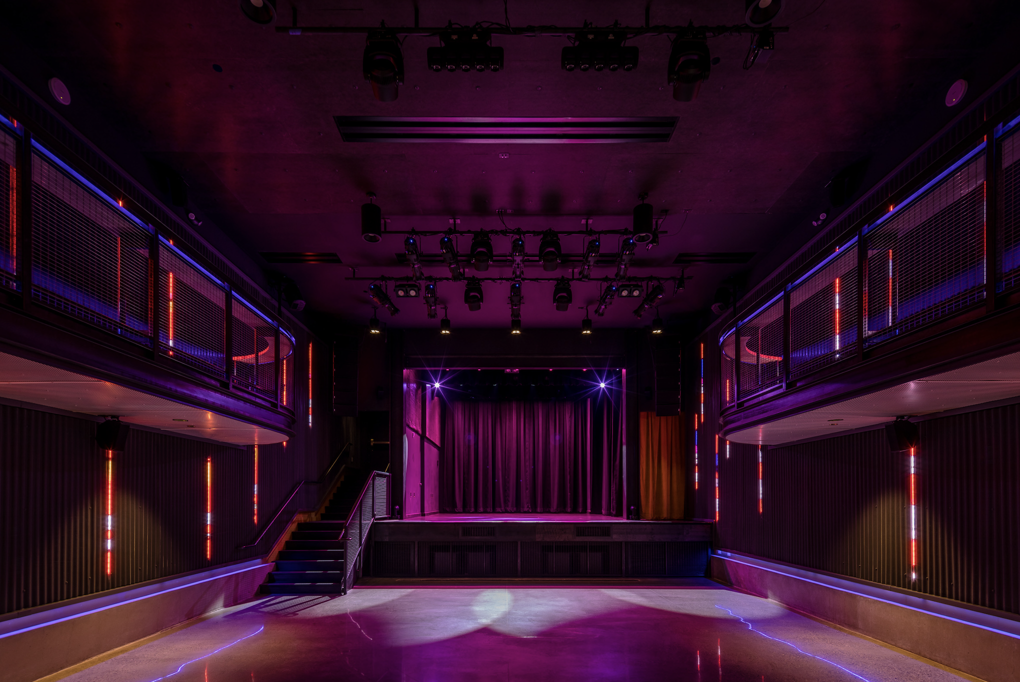 Empty stage with purple curtains and overhead stage lights in a nightclub or theater with purple and red neon lighting on the walls.
